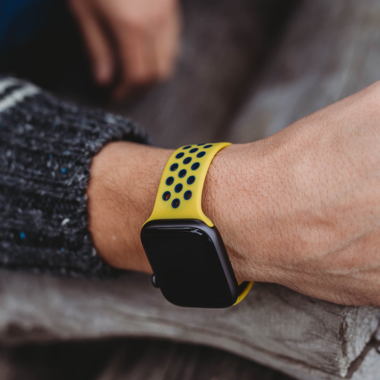 Die Person trägt ein arktisband Apple Watch Silikonarmband „Energy“ – schwarz mit gelben, schwarz gepunkteten Silikonstreifen – und ruht mit dem Arm auf einer Holzoberfläche. Der dunkle Pullover harmoniert perfekt mit dem stylischen Sport-Accessoire von arktisband.