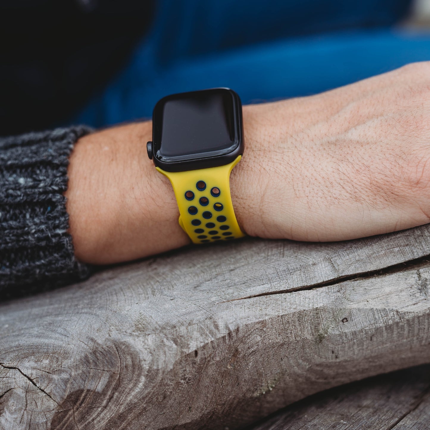 Die Person trägt ein arktisband Apple Watch Silikonarmband „Energy“ in Gelb und legt ihren Arm auf eine verwitterte Holzoberfläche. Das moderne arktisband Accessoire sticht durch den dunklen, strukturierten Pullover hervor.