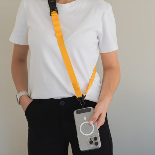 Eine Person in weißem T-Shirt und schwarzer Hose hält ein Smartphone mit grauer Hülle, befestigt am gelben arktis Smartphone Gurtband Lanyard von arktis, das sie crossbody mit 1-Klick-Verschluss trägt; die andere Hand steckt in der Tasche.