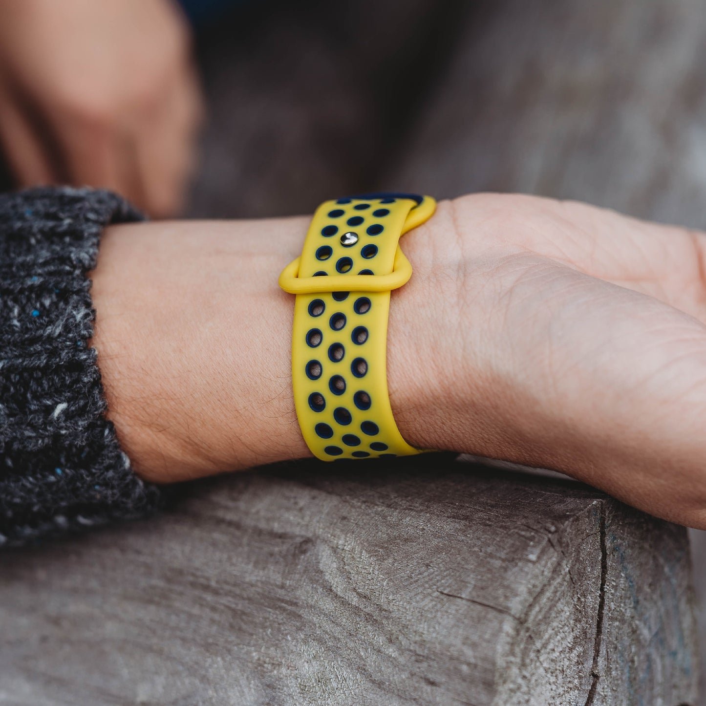 Eine Nahaufnahme eines Handgelenks mit dem Arktisband Apple Watch Silikonarmband „Energy“ in Gelb und Schwarz; das perforierte Sportarmband liegt auf einer Holzoberfläche, eine dunkle strukturierte Hülle ist sichtbar.