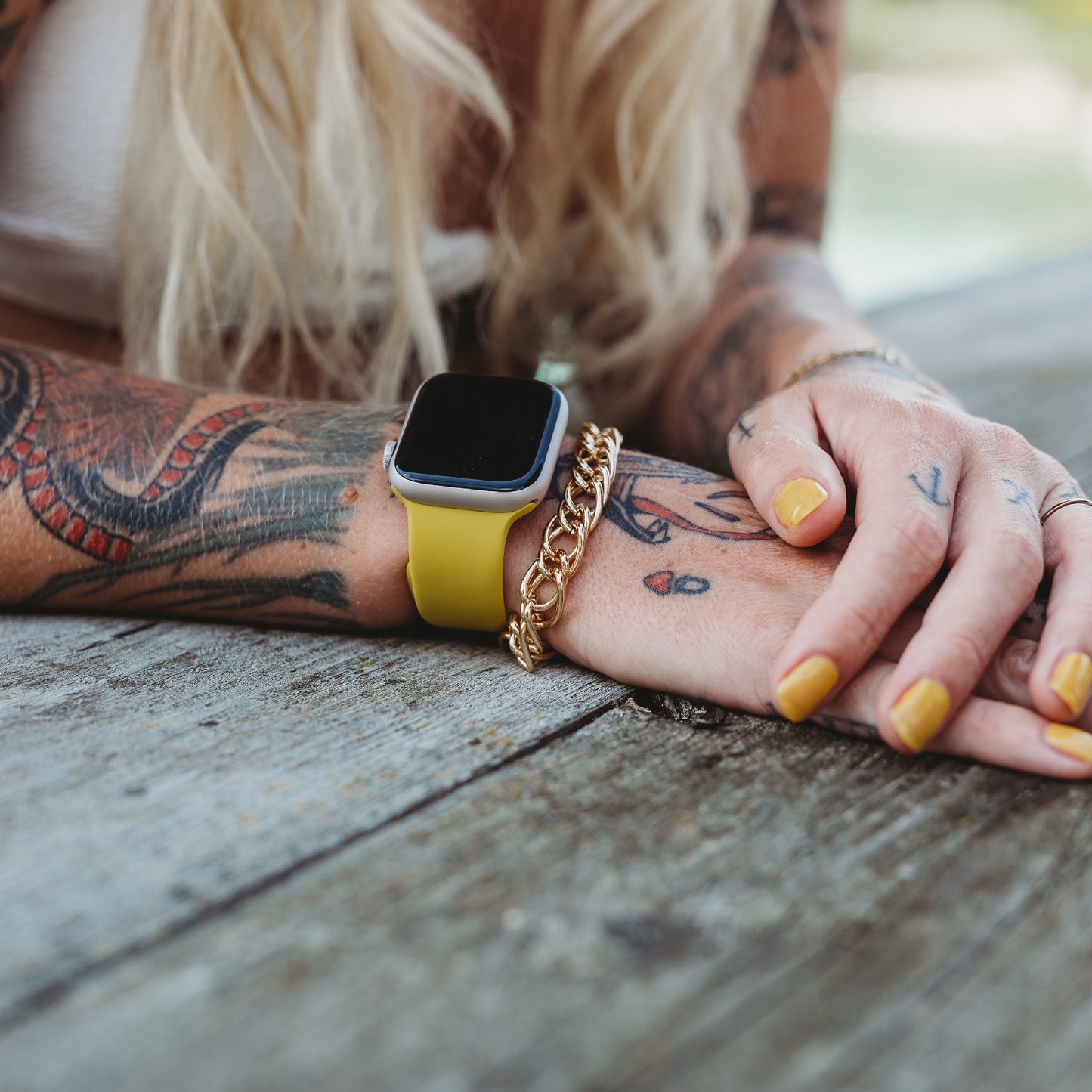 Eine blonde Person mit Tattoos legt ihre Hände auf Holz und trägt ein Arktisband Apple Watch Silikonarmband „Aqua“, ein goldenes Kettenarmband und gelben Nagellack für einen abgestimmten, sportlichen Stil.