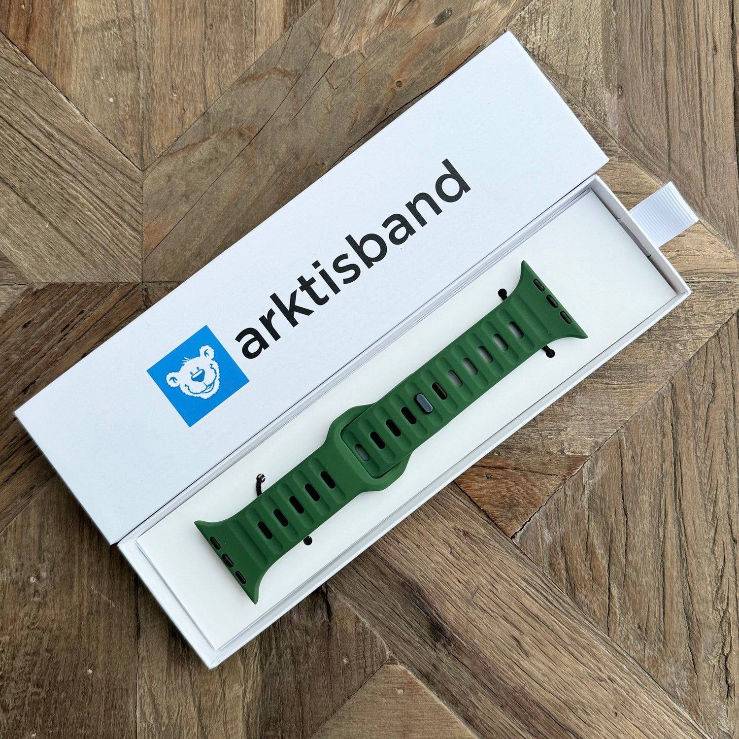 Ein grünes Arktisband-Silikonarmband „Active“ für die Apple Watch mit atmungsaktivem Design wird in einer weißen Box mit blauem Arktisband-Branding gezeigt und ruht auf einer Holzoberfläche mit Fischgrätenmuster.