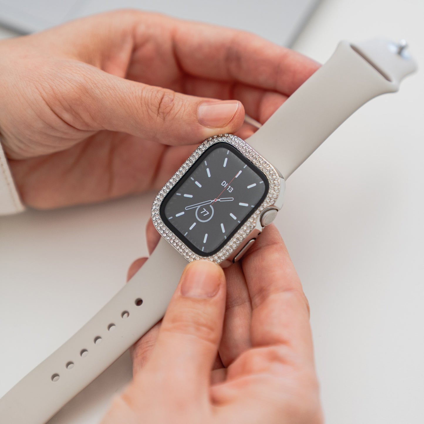 Eine Person hält die arktisband GLAMOUR Glitzerhülle für Apple Watch mit weißem Band, strassbesetzter Lünette und gehärtetem Schutzglas über einem minimalistischen analogen Zifferblatt.