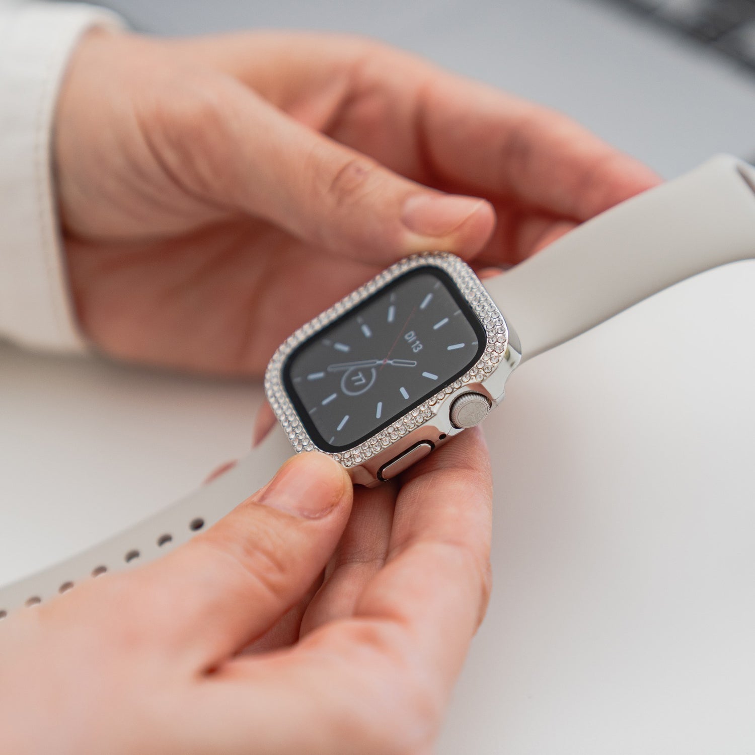 Eine Person hält eine Apple Watch mit weißem Armband und einer Arktisband GLAMOUR Glitzerhülle für Apple Watch mit gehärtetem Schutzglas. Das analoge Zifferblatt ist sichtbar, im Hintergrund befindet sich eine unscharfe Tastatur.