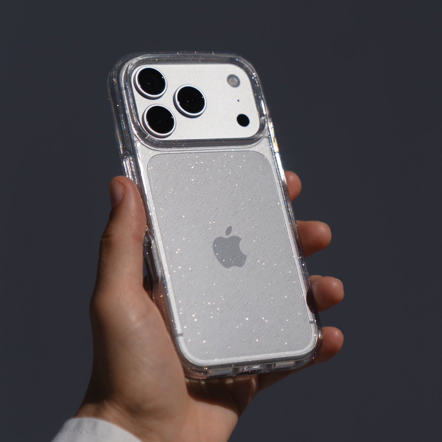 Eine Hand hält das arktis iPhone 17 Pro Max Glitter Case, eine transparente Schutzhülle von arktis, auf einem weißen iPhone vor einem schlichten dunklen Hintergrund.