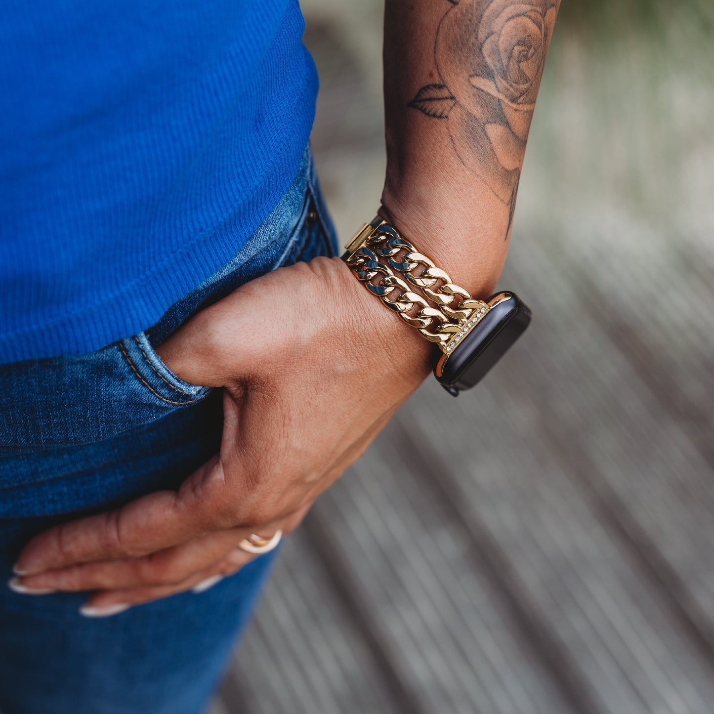 Diese Person trägt das arktisband Apple Watch Armband „Princess“ von arktisband, eine Smartwatch mit goldener Kette und glanzvoller Gliederstruktur, und kombiniert es mit Bluejeans, einem blauen Oberteil, einem Rosentattoo auf dem Unterarm und einem Ring.