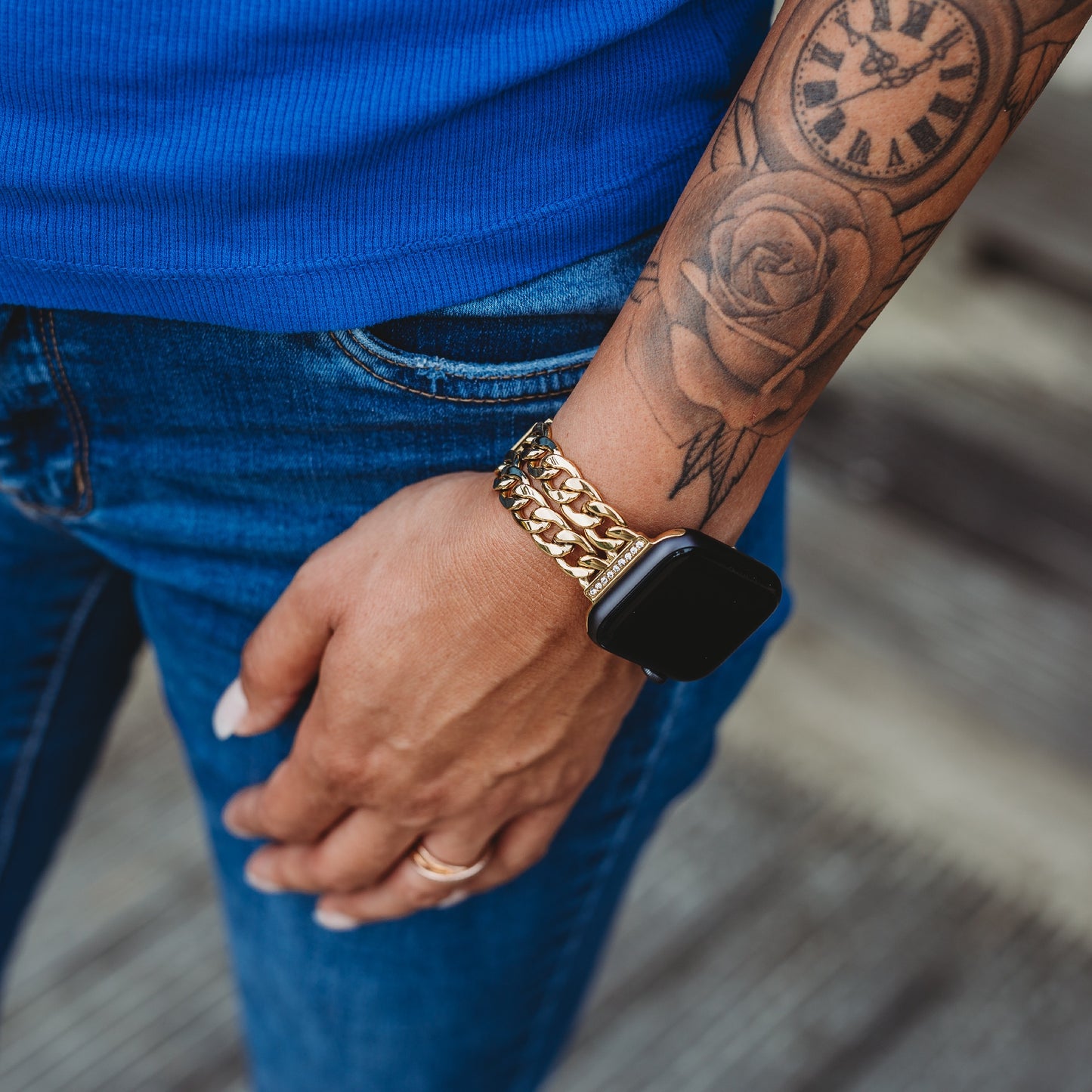 Eine Person in Blue Jeans und blauem Hemd trägt das arktisband Apple Watch Armband „Princess“ von arktisband mit glanzvoller Gliederstruktur auf einem goldenen Kettenband, auf ihrem Unterarm sind eine Uhr und ein Rosentattoo sichtbar.