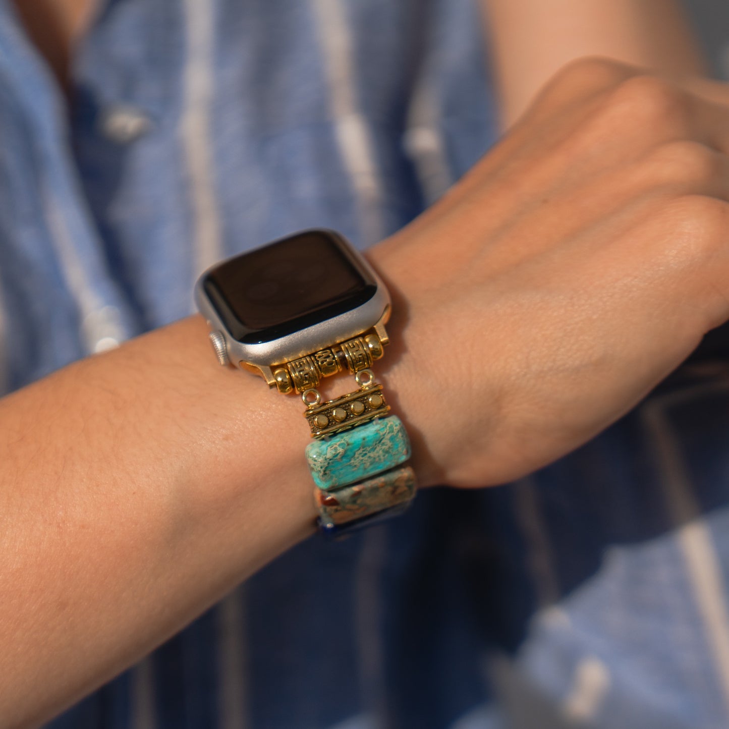 Eine Person trägt ein Arktisband Apple Watch „Stone Loop“-Armband – ein mit Perlen besetztes Armband aus türkisfarbenem und goldenem Naturstein – für ihre Apple Watch am Handgelenk.
