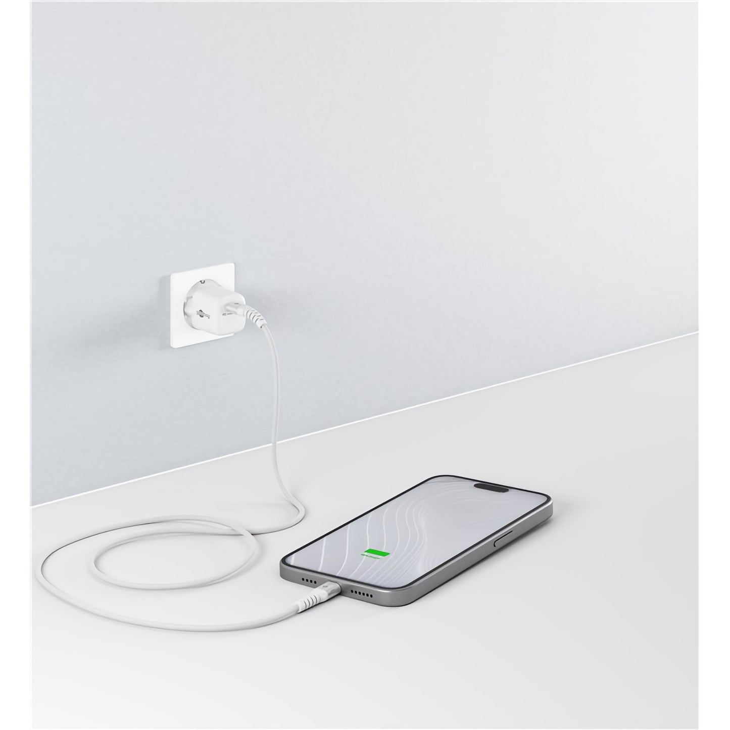 Ein Smartphone liegt flach auf weißer Oberfläche und lädt per Goobay USB-C auf USB-C Textilkabel mit Metallsteckern, das an einer hellen Wandsteckdose angeschlossen ist. Das Display zeigt das Ladesymbol.
