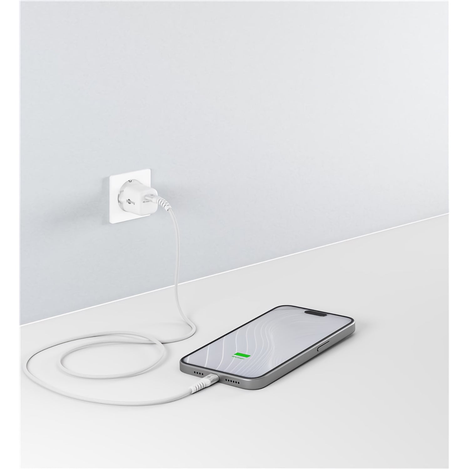 Ein Smartphone liegt flach auf weißer Oberfläche und lädt per Goobay USB-C auf USB-C Textilkabel mit Metallsteckern, das an einer hellen Wandsteckdose angeschlossen ist. Das Display zeigt das Ladesymbol.