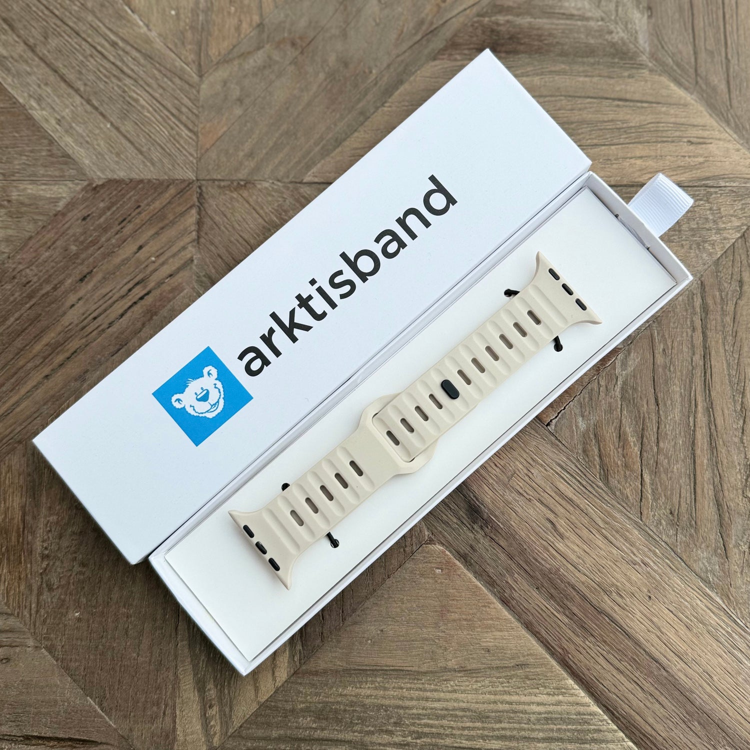 Ein hellbeiges Arktisband Apple Watch Silikonarmband „Active“ wird in einer weißen Box mit dem Arktisband-Logo und einem Eisbärsymbol auf dem Deckel gezeigt und ruht auf einer Holzoberfläche.