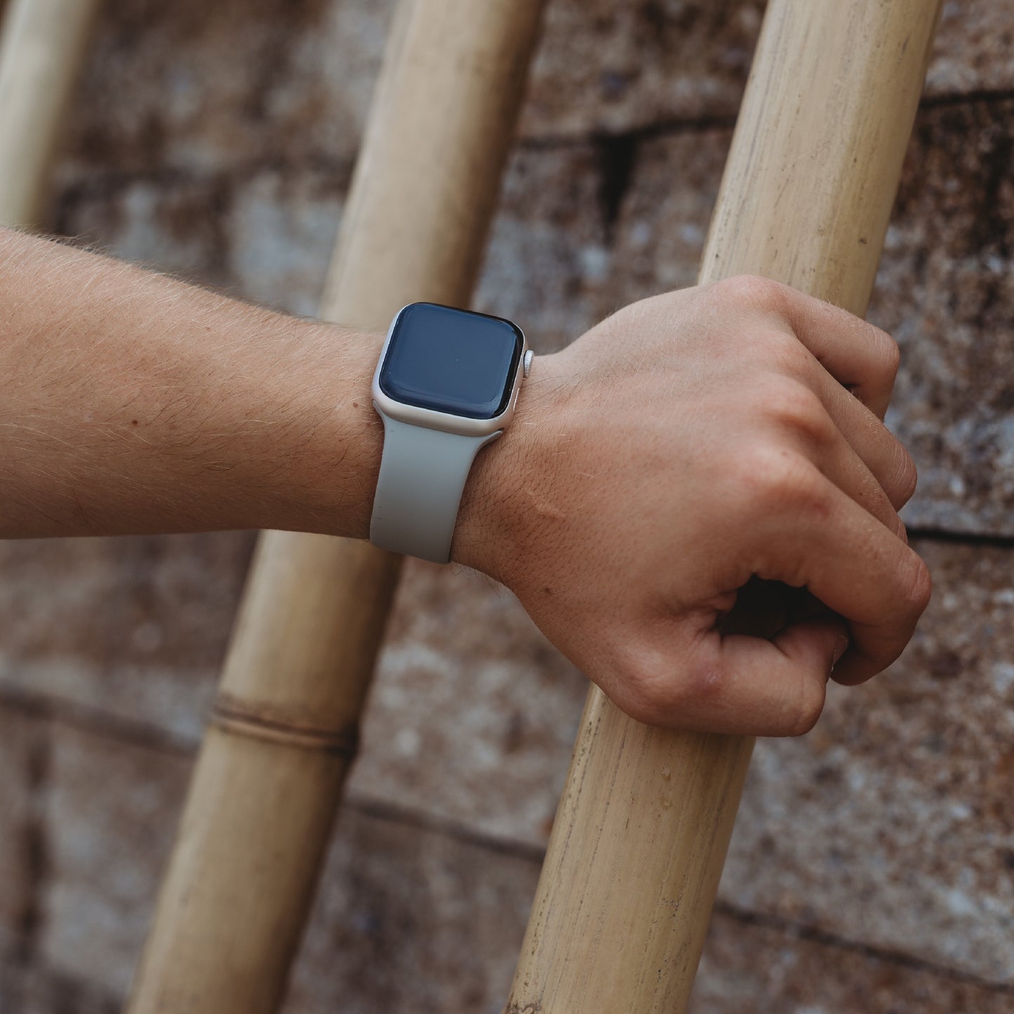 Eine Person, die das arktisband Apple Watch Silikonarmband „Aqua“ mit hellgrauem Band trägt, stützt ihren Arm auf Bambusstangen an einer Steinmauer ab und zeigt, wie das sportliche arktisband Accessoire jeden lässigen Stil ergänzt und perfekt zur Apple Watch passt.