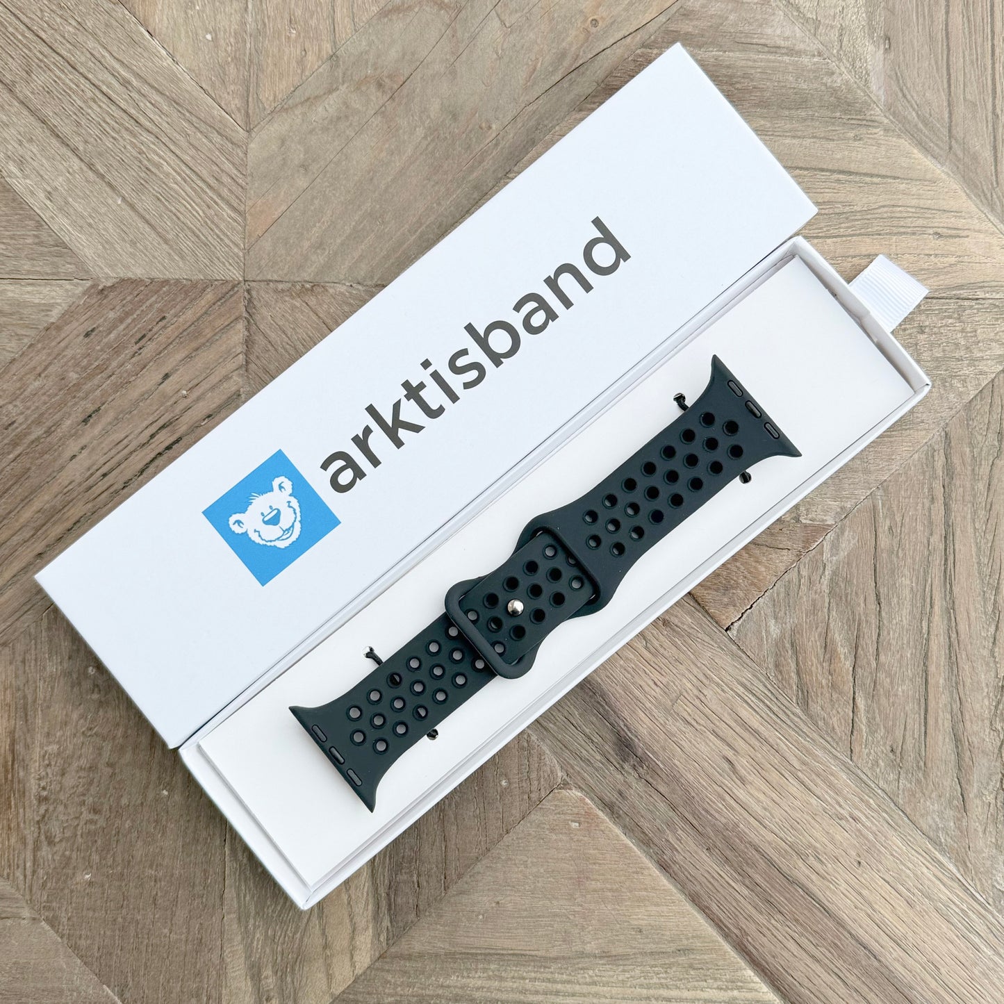 Das arktisband Apple Watch Silikonarmband „Energy“ von arktisband wird in Schwarz mit Perforationen gezeigt und in einer offenen weißen Box mit einem blauen Eisbär-Logo auf einer Holzoberfläche mit Chevron-Muster präsentiert – ideal fürs Training oder zum täglichen Tragen.