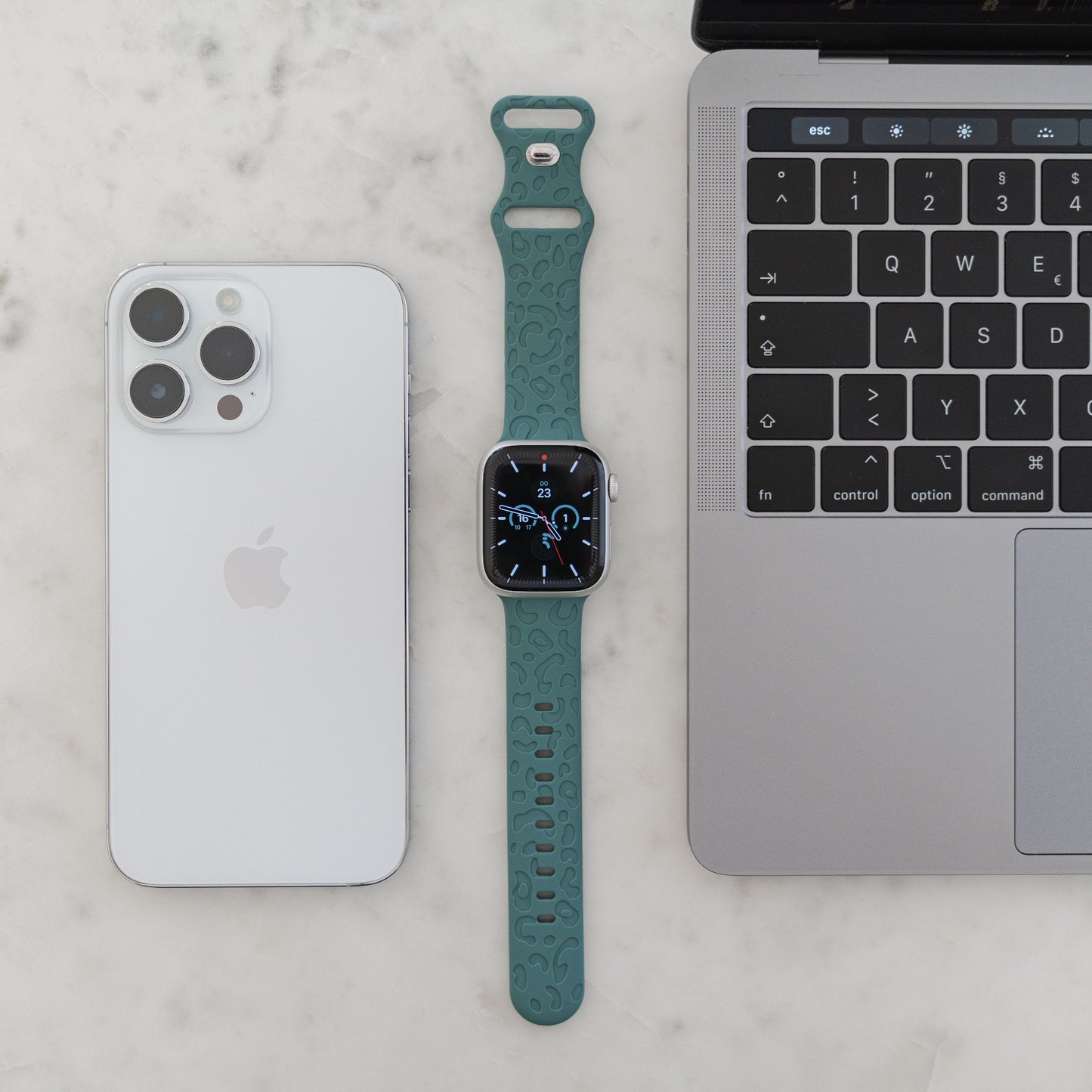 Ein silbernes iPhone, ein Arktisband Apple Watch Armband „Leopard“ in Blaugrün auf einer kompatiblen Smartwatch und ein silberner Laptop mit schwarzer Tastatur sind ordentlich auf einer weißen Marmoroberfläche angeordnet.