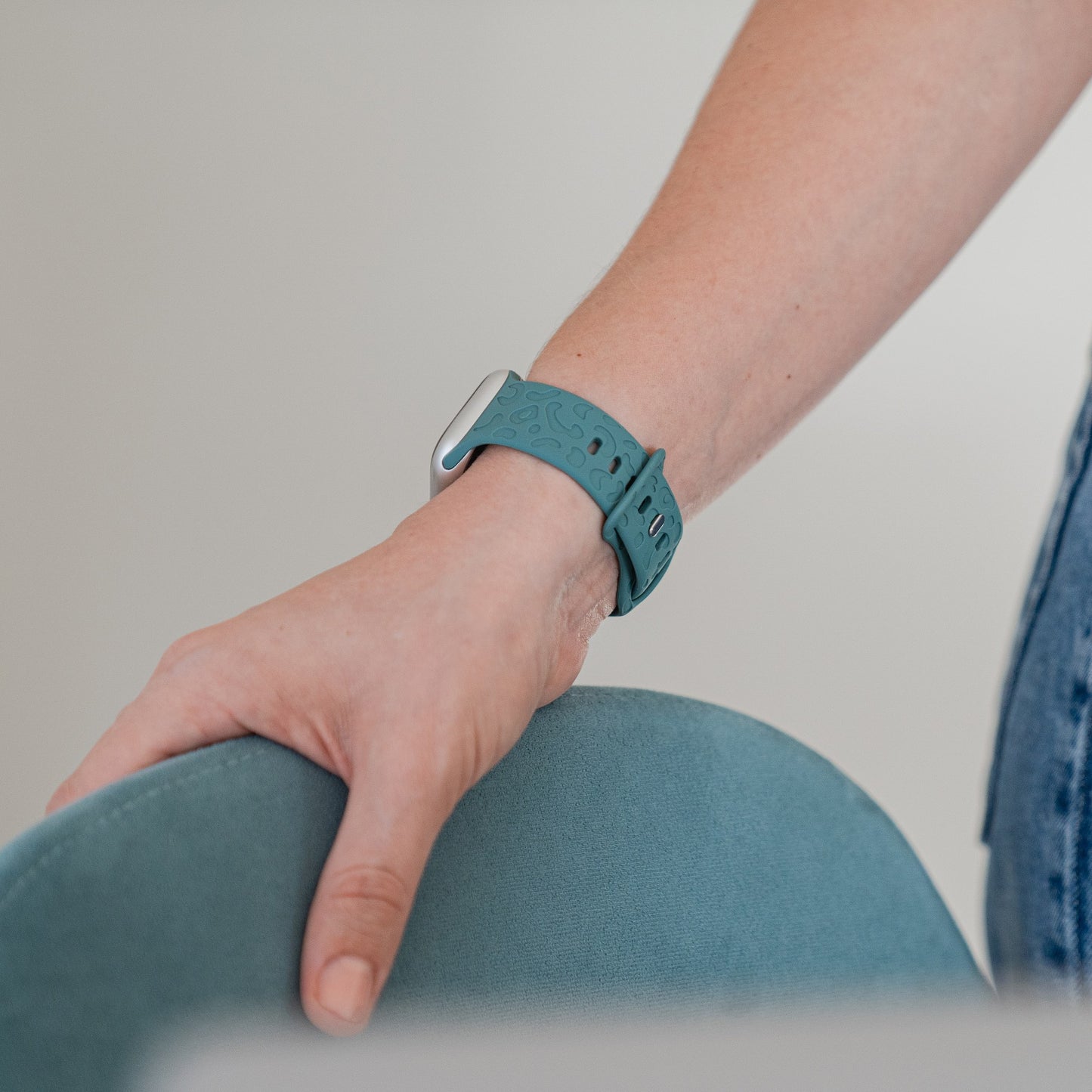 Jemand mit dem Arktisband Apple Watch Armband „Leopard“ klammert sich an die Rückenlehne eines hellblauen Stuhls. Vor einem neutralen Hintergrund sind lediglich sein Arm und ein Teil seines Oberkörpers in Freizeitkleidung zu sehen.