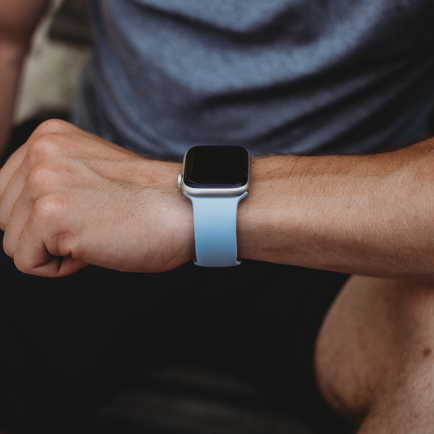 Eine Person trägt ein Arktisband Apple Watch Silikonarmband „Aqua“ am linken Handgelenk, dazu ein graues Shirt und dunkle Shorts. Der Arm liegt auf dem Bein. Der Bildschirm der Uhr ist ausgeschaltet.