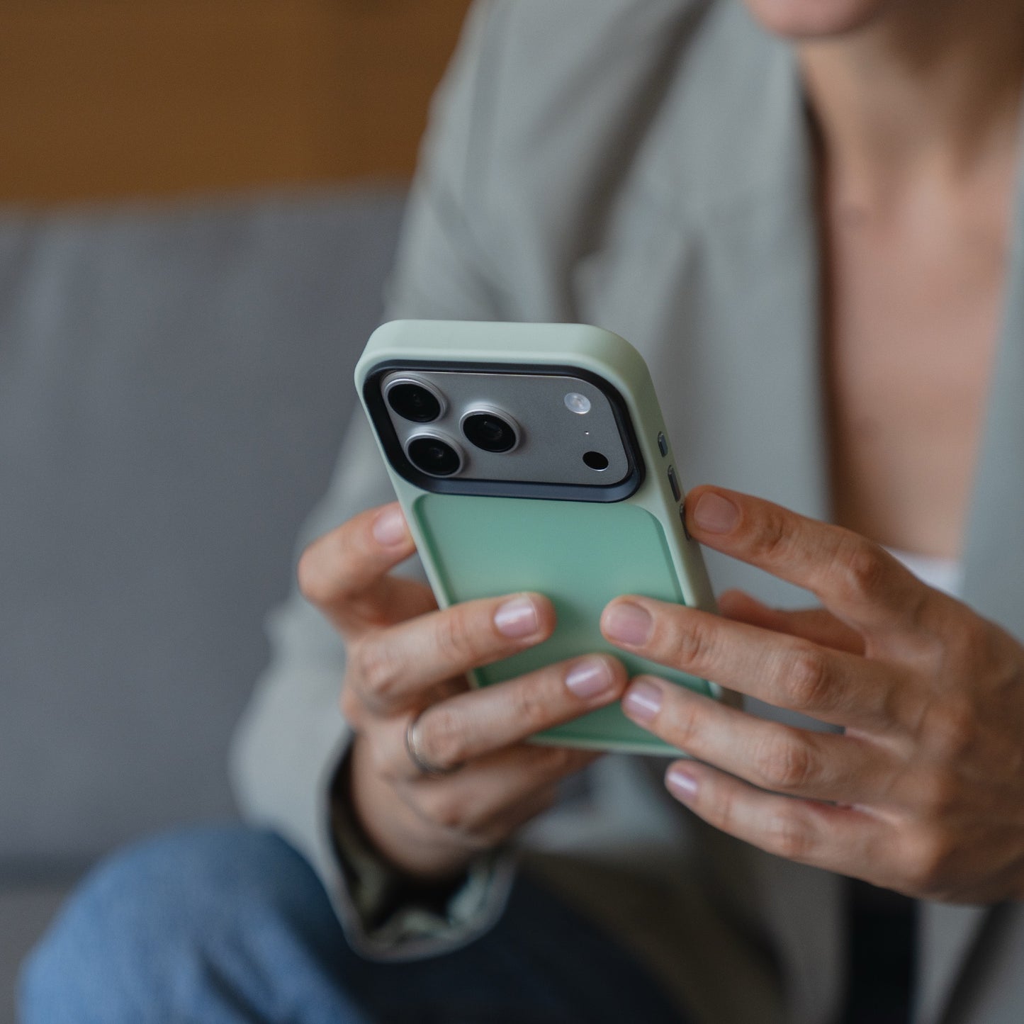 Eine Person in einem Blazer hält ein Smartphone mit einem Arktis iPhone 17 Pro Solid Case in mattem Grün, während sie auf einer Couch sitzt.