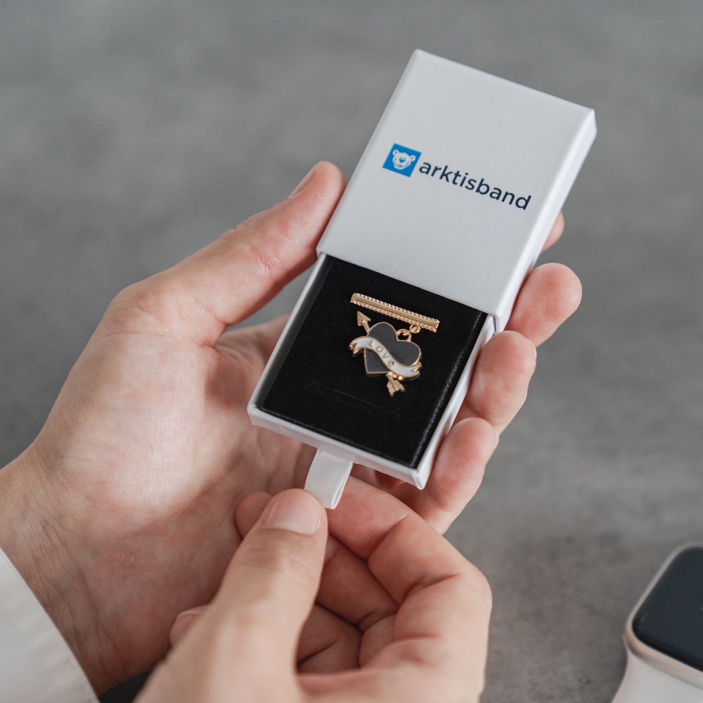 Eine Person hält eine offene weiße Arktisband-Box mit einem Apple Watch Armband Charm – einer Anstecknadel mit Herz und Banner auf schwarzem Samt – von Arktisband, ideal als individuell anpassbares Accessoire.