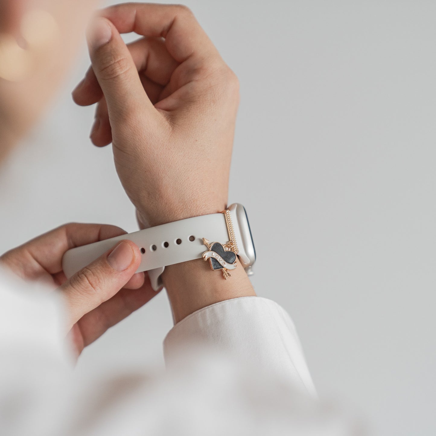 Eine Hand befestigt ein weißes arktisband Apple Watch Armband von arktisband, kombiniert mit einem goldenen Armband mit Vogelanhänger. Der helle Hintergrund unterstreicht das individuell gestaltbare Design und den anpassbaren Stil.