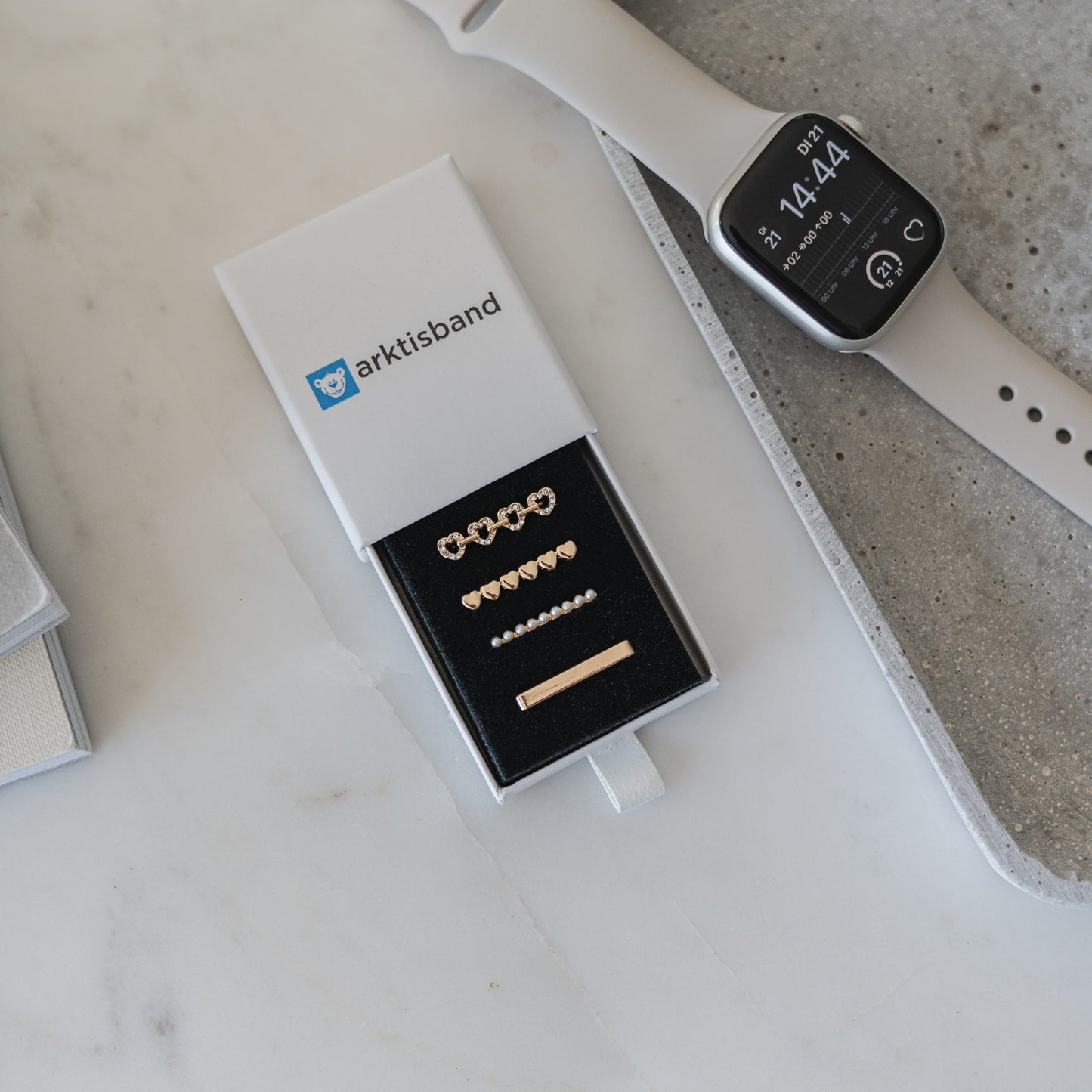 Eine Schachtel mit der Aufschrift „arktisband“ und sechs goldenen „arktisband Apple Watch Armband Charms“ liegt auf einer Marmoroberfläche neben einer beigen Smartwatch und einem grauen Tablett – perfekt, um dein Apple Watch Armband individuell zu gestalten.