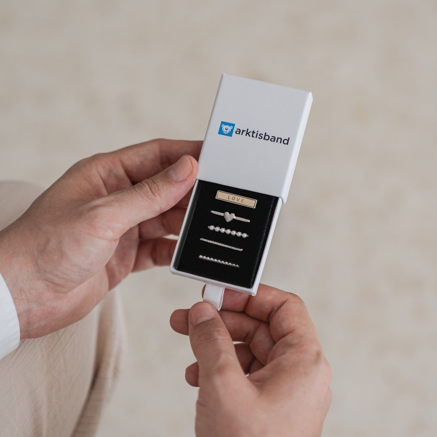 Eine Person hält eine offene weiße Arktisband-Box mit fünf metallischen Arktisband-Apple-Watch-Armbandanhängern, darunter einer mit der Aufschrift „LOVE“ und einem Herz, perfekt zum Personalisieren Ihres Apple-Watch-Armbands.