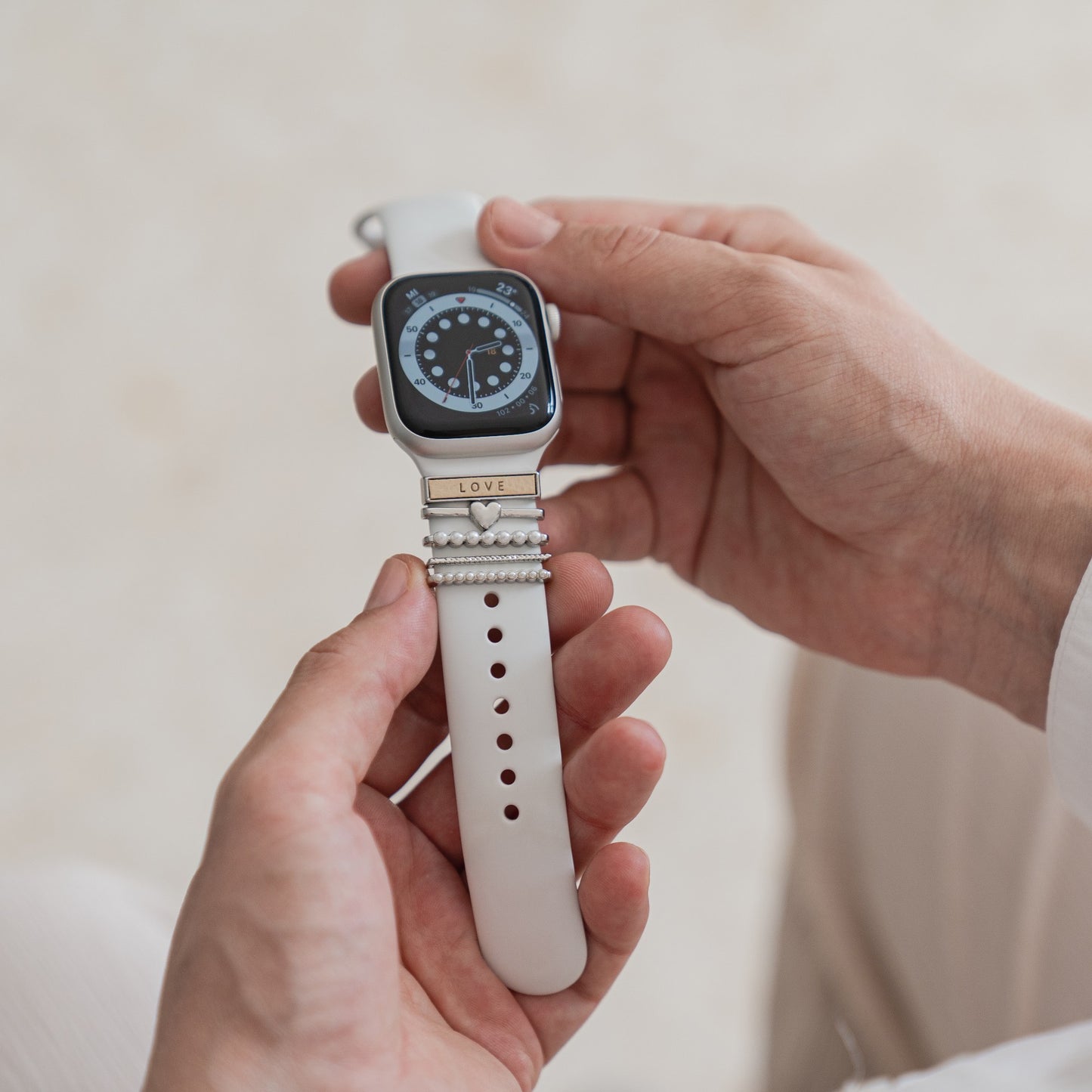 Eine Person hält eine arktisband Apple Watch mit weißem Armband, verziert mit den arktisband „Love“ Apple Watch Charms in Silber. Der Charm besteht aus Edelstahl und Glas und trägt die Gravur „LOVE“; das Display zeigt eine analoge Uhr.