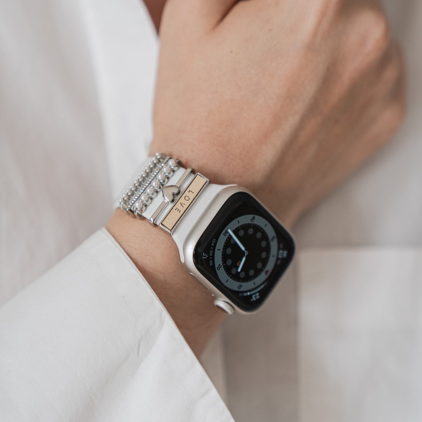 Eine Person trägt eine silberne Apple Watch mit arktisband Armband-Charms – Perlen und einem metallischen LOVE-Akzent – und ein weißes Langarmshirt. Der Stil lässt sich mit arktisband Apple Watch Armband-Charms von arktisband individuell gestalten.