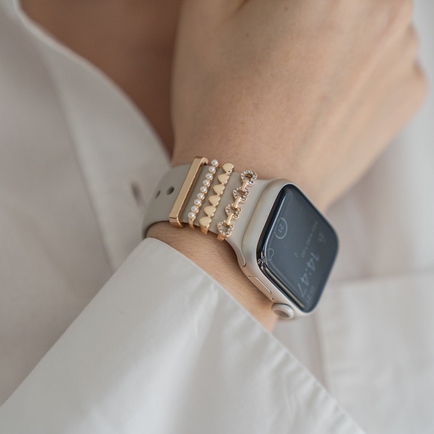 Eine Person trägt ein weißes Langarmshirt und ein Arktisband Apple Watch Armband mit vier goldenen und juwelenbesetzten Arktisband Apple Watch Armband-Anhängern. Auf dem Smartwatch-Bildschirm werden die Uhrzeit und weitere Daten angezeigt.