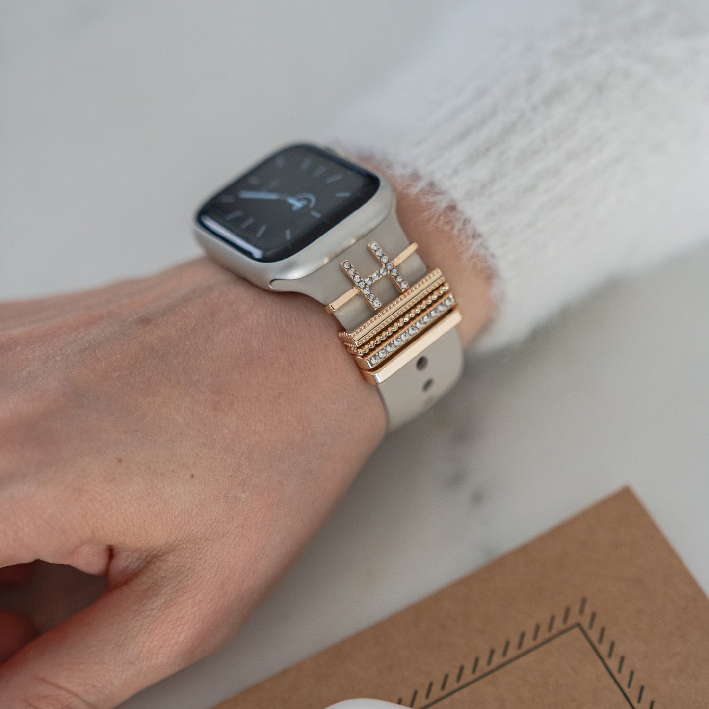 Eine Hand trägt eine Smartwatch mit goldenen und silbernen Apple Watch-Charms im „H-Stil“ von arktisband an einem hellen Armband, was für dekoratives Flair sorgt.
