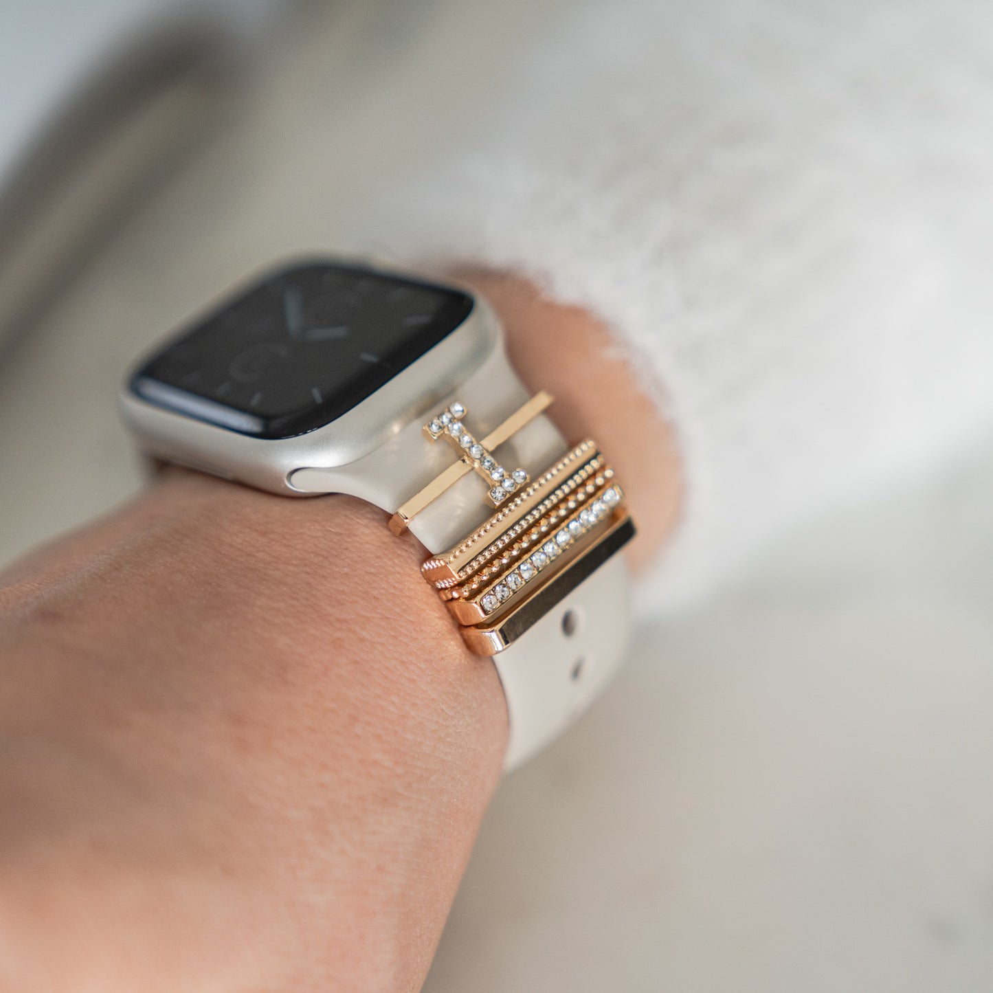 Eine Smartwatch mit leichtem Arktisband ist mit den Arktisband Apple Watch Charms „I Style“ in Gold und Silber gestaltet, akzentuiert durch kleine klare Steine. Das Handgelenk ist mit einer leichten Strickmanschette versehen.