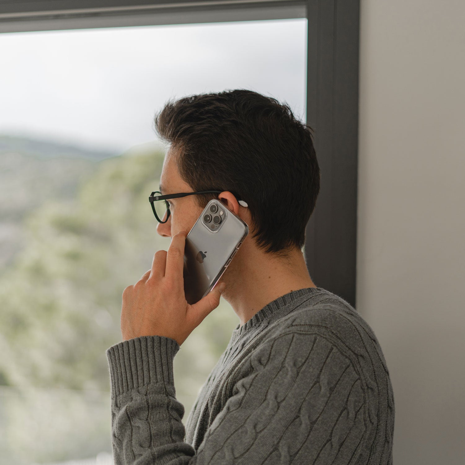 Eine Person mit Brille und grauem Pullover hält ein iPhone mit dem arktis Invisible Air Case von arktis ans Ohr und telefoniert, während sie im Innenbereich neben einem Fenster steht.