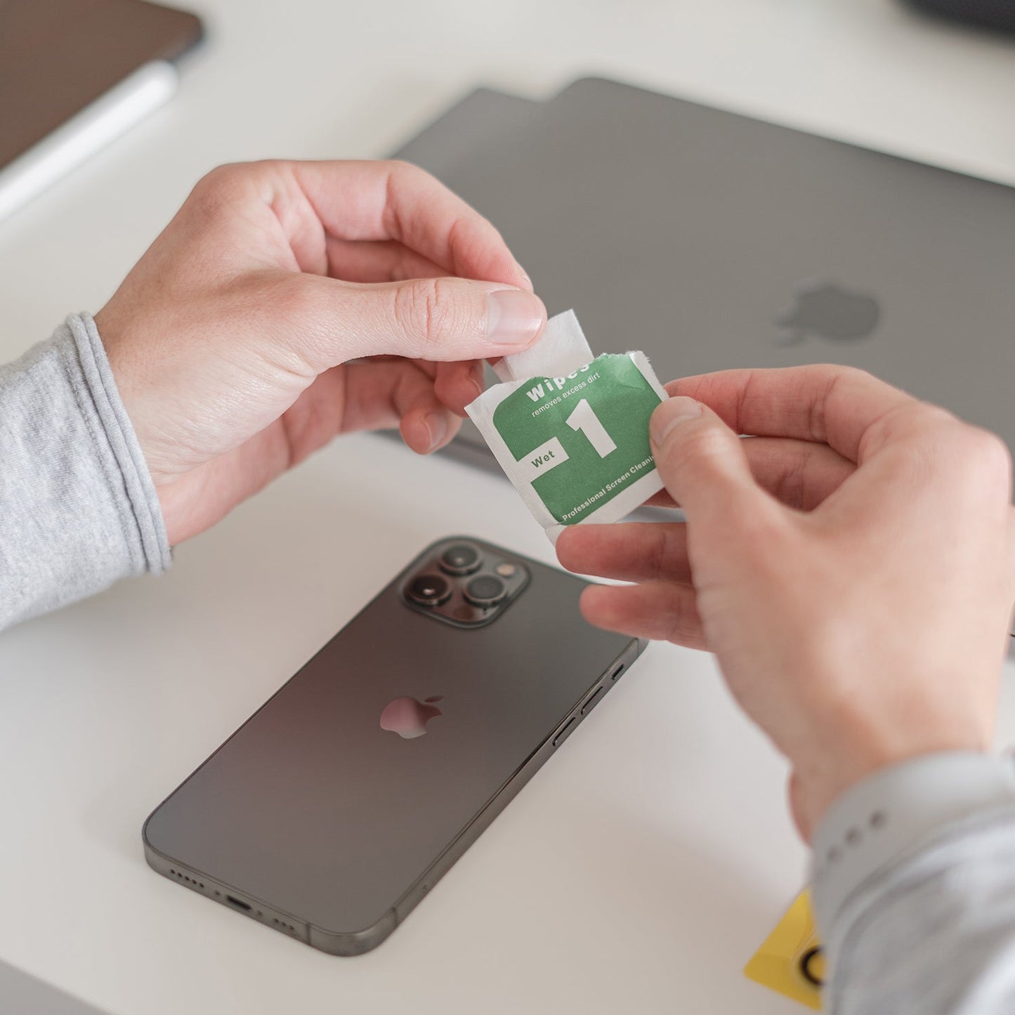 Hände halten ein Reinigungstuch über ein iPhone und einen geschlossenen Laptop auf einem weißen Schreibtisch und bereiten das Gerät für den arktis iPhone Air Kamera Protector von arktis vor.