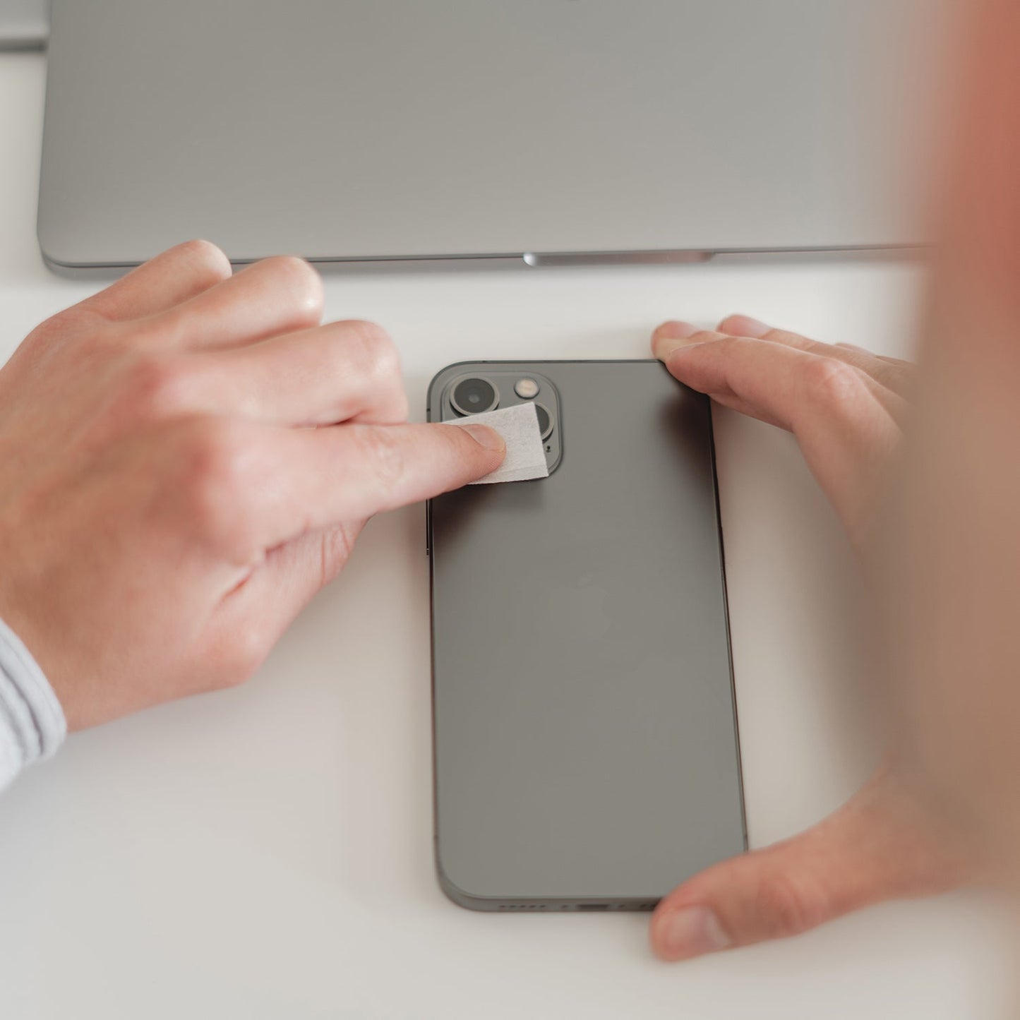 Eine Person wischt ihr Smartphone in der Nähe eines Laptops ab. So bleibt der arktis iPhone Air Kamera Protector von arktis sauber und bietet optimalen Schutz.