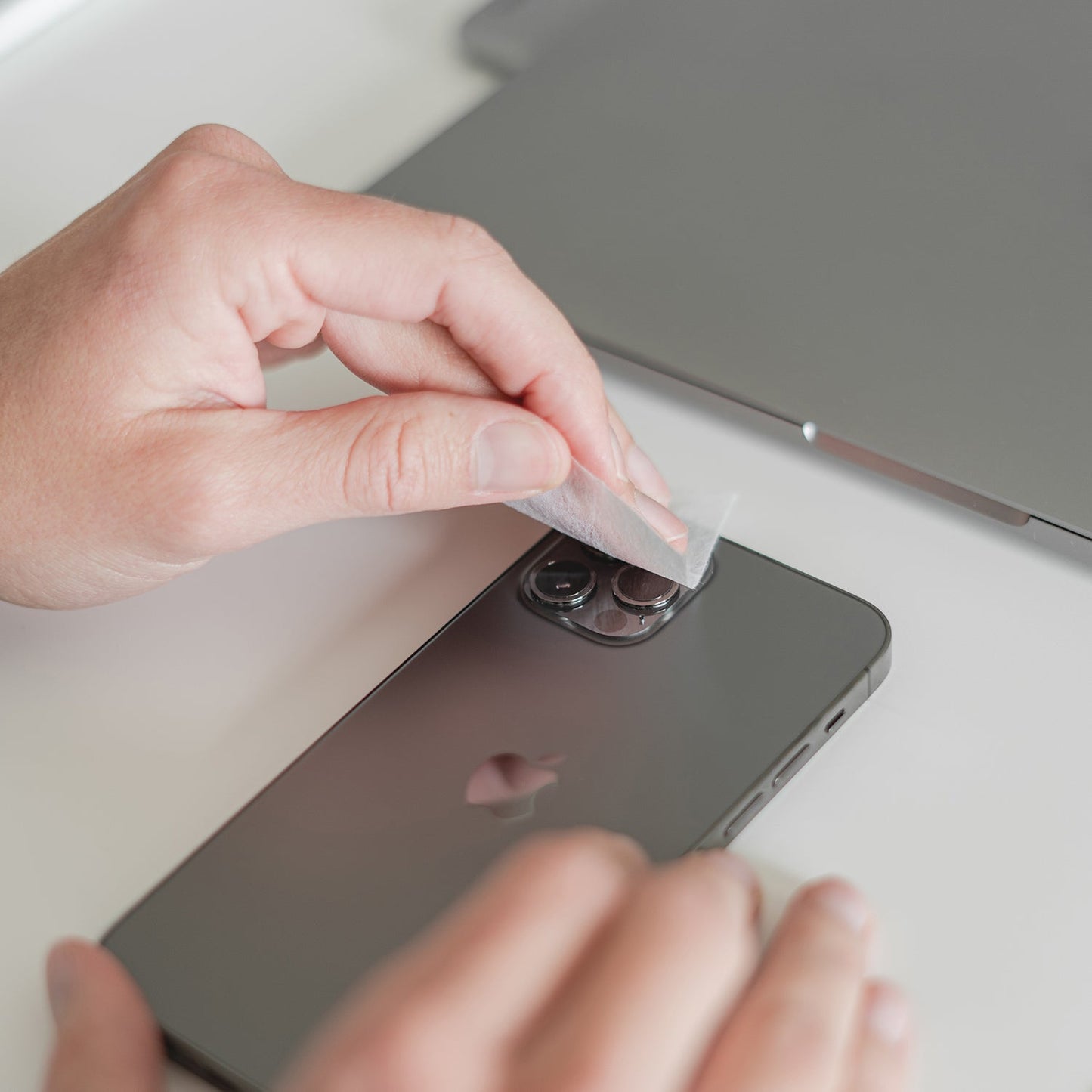 Jemand zieht die Schutzfolie von einem arktis iPhone Air Kamera Protector von arktis ab, der neben einem geschlossenen Laptop auf einem Schreibtisch steht.
