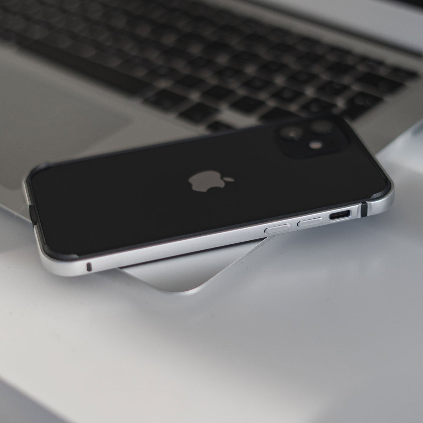 Ein arktis iPhone Air Bumper in Schwarz aus stoßabsorbierendem TPU-Silikon liegt mit der Vorderseite nach unten auf einer weißen Fläche neben einer silbernen Laptoptastatur.