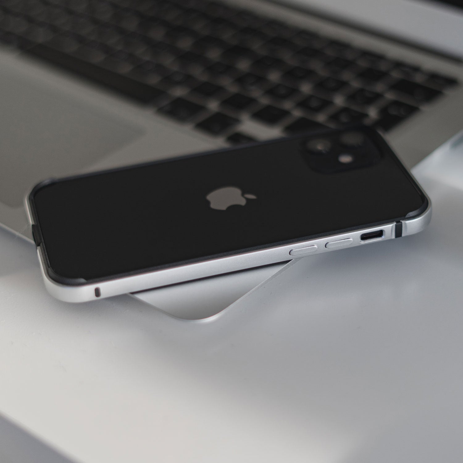 Ein arktis iPhone Air Bumper in Schwarz aus stoßabsorbierendem TPU-Silikon liegt mit der Vorderseite nach unten auf einer weißen Fläche neben einer silbernen Laptoptastatur.