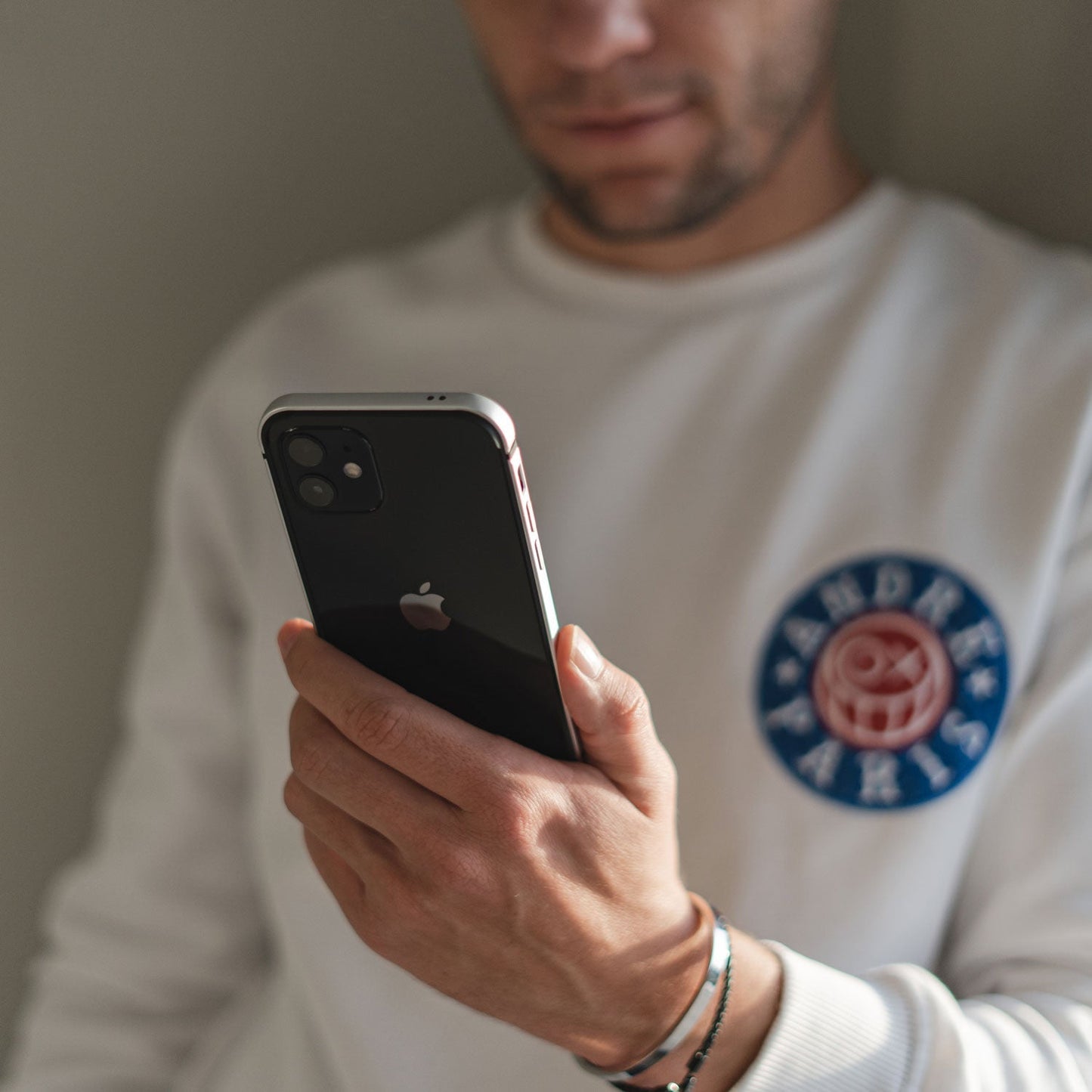 Eine Person in einem weißen Sweatshirt hält und betrachtet ein schwarzes iPhone, geschützt durch den arktis iPhone Air Bumper von arktis, der aus ultraleichtem, stoßabsorbierendem TPU-Silikon besteht und für das iPhone 17 geeignet ist.