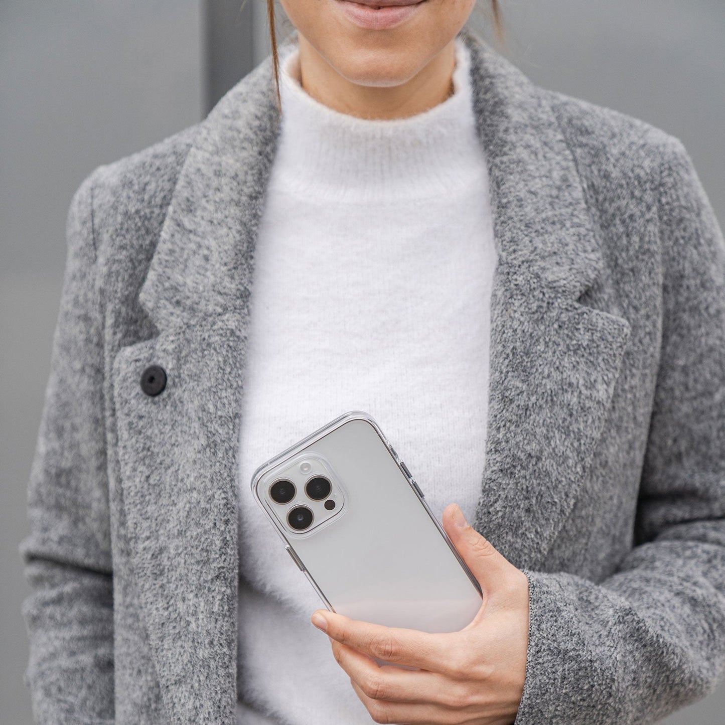 Eine Frau im grauen Mantel hält ein iPhone 15 Pro mit der MR PROTECT CLEAR-Hülle und steht vor einem neutralen Hintergrund.