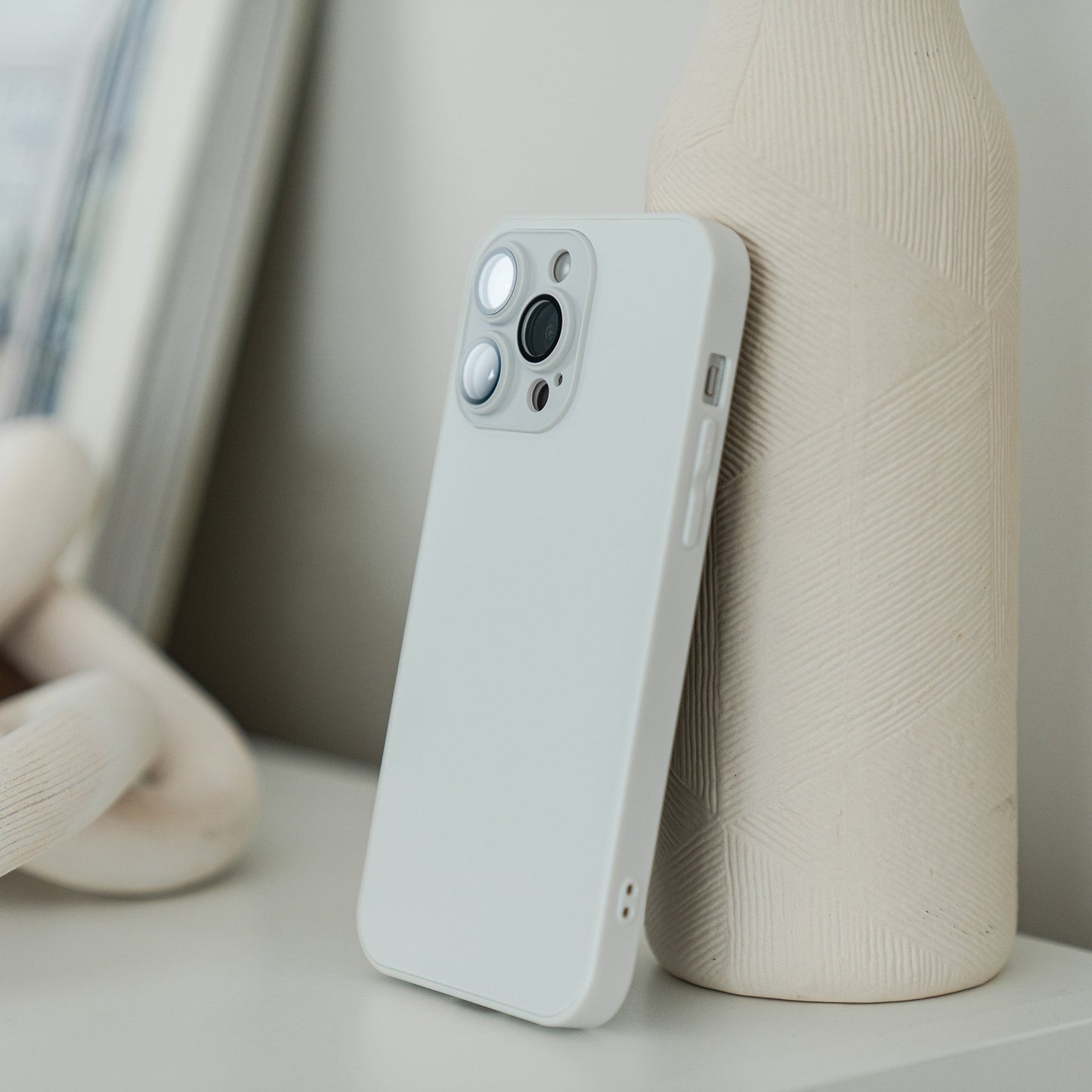 An arktis iPhone 16e Tech Case in white props up a smartphone against a textured, light-colored ceramic vase on a white surface, with a blurred picture frame visible in the background.