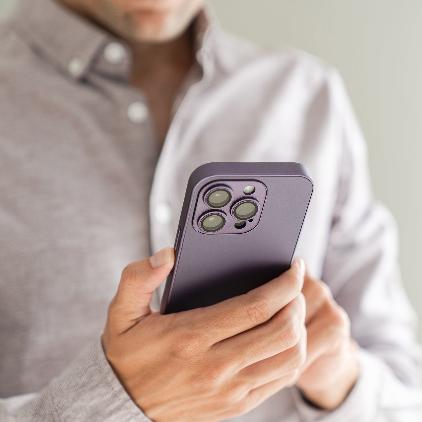 Eine Person in einem hellgrauen Hemd hält ein Smartphone mit dem arktis iPhone 16 Tech Case von arktis – einer violetten, stoßabsorbierenden TPU-Silikon-Hülle – und fokussiert die Kameralinsen des Telefons. Das Gesicht der Person ist teilweise außerhalb des Bildes.