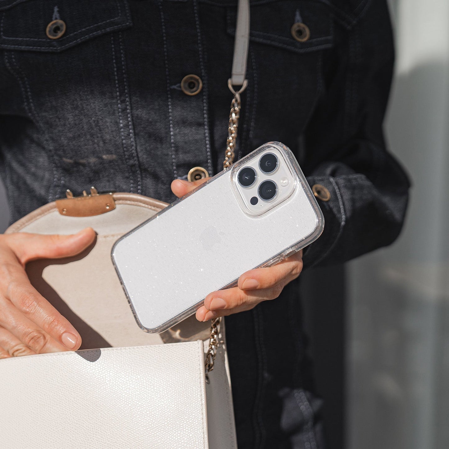 Eine Person in einer dunklen Jeansjacke hält in der einen Hand ein weißes Smartphone mit einem Arktis iPhone 16e Glitter Case und in der anderen eine helle Handtasche.
