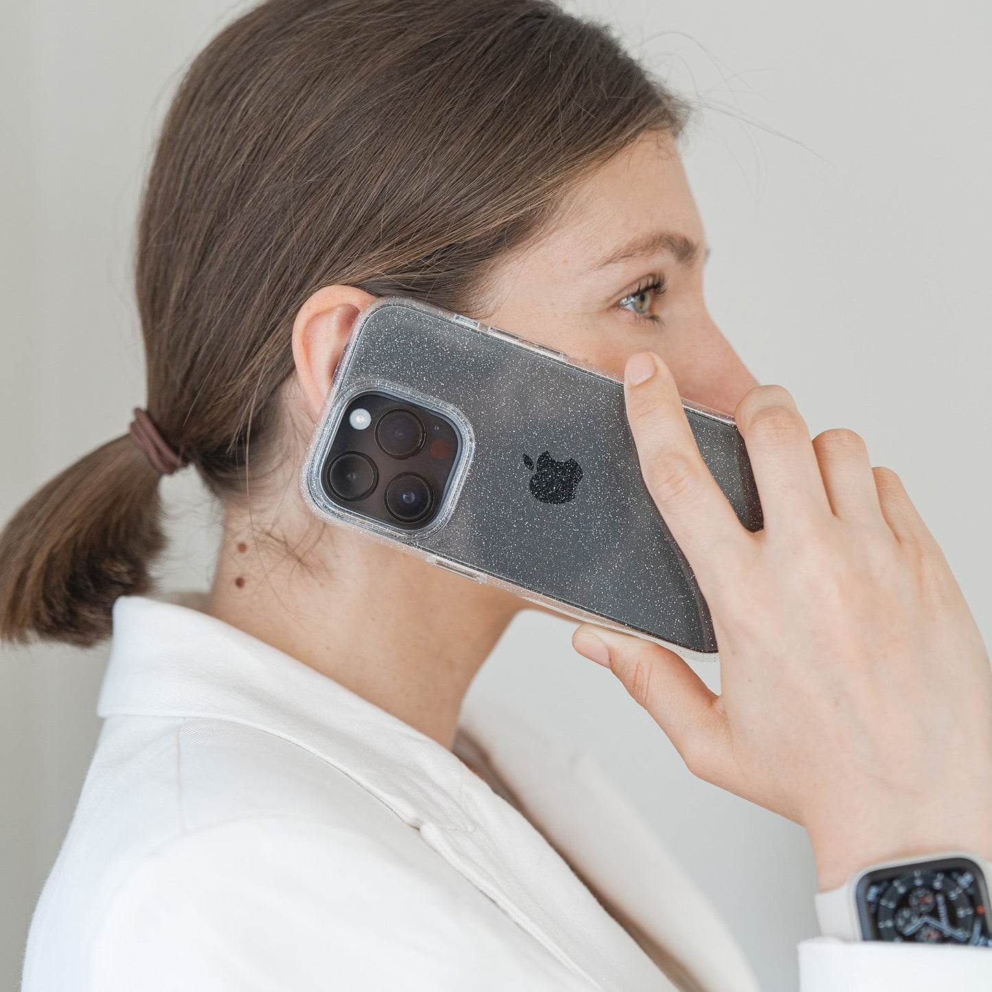 Eine Person mit braunen Haaren zu einem tiefen Pferdeschwanz hält ein schwarzes iPhone in einem arktis iPhone 16e Glitter Case von arktis ans Ohr. Sie trägt einen weißen Blazer und eine Smartwatch und steht vor einem neutralen Hintergrund.