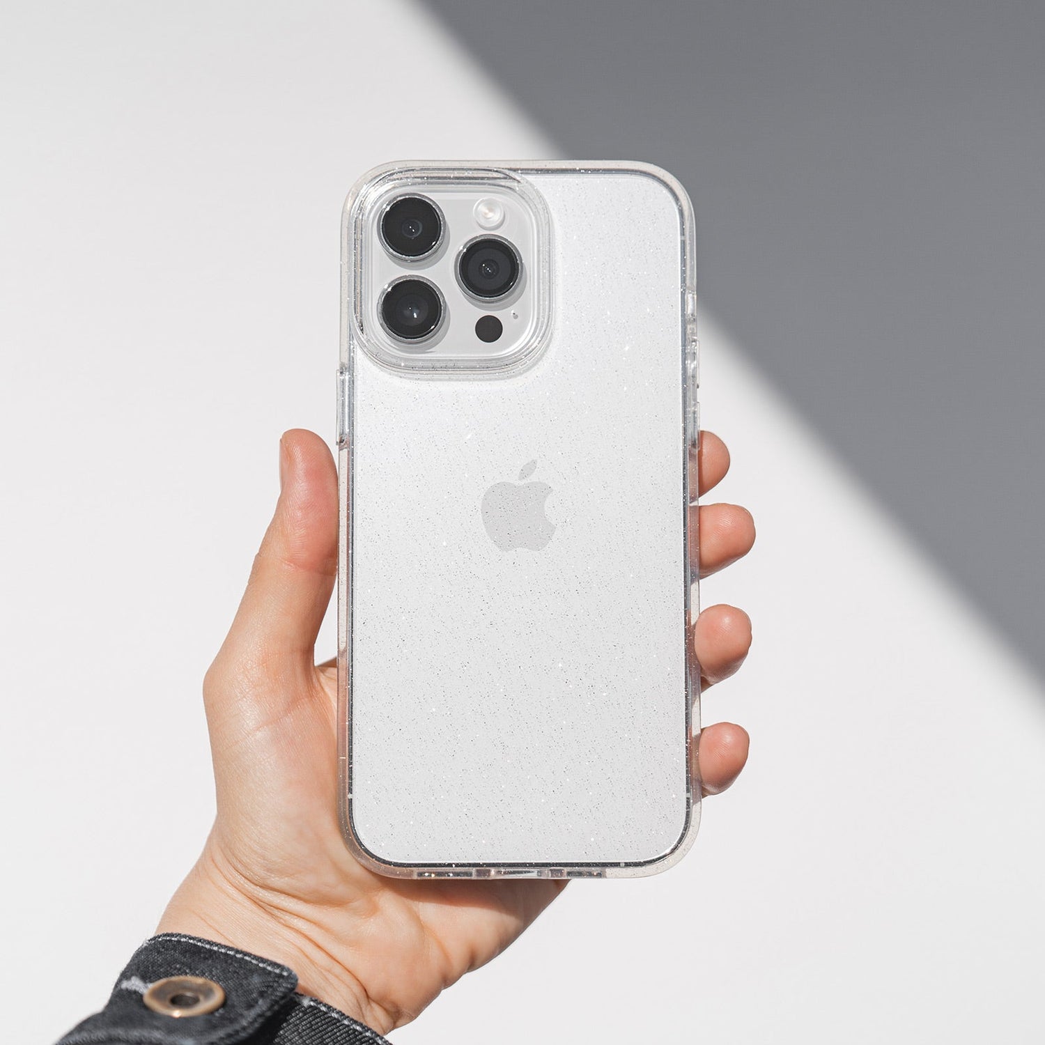 Eine Hand hält ein arktis iPhone 16e Glitter Case mit Apple-Logo und Dreifachkamera. Das transparente TPU-Silikon-Case von arktis funkelt im Licht.