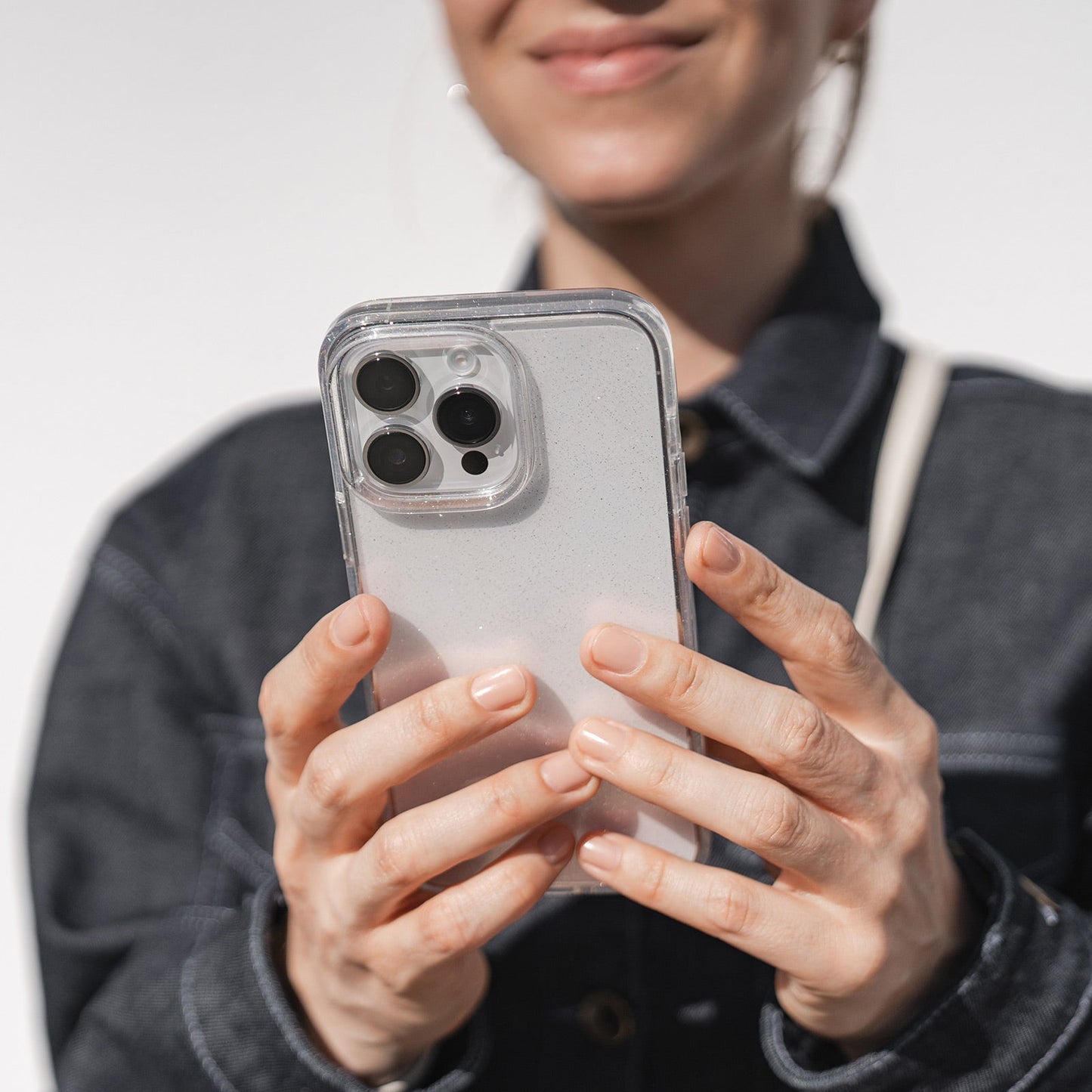 Eine Person in einer dunklen Jeansjacke hält das arktis iPhone 16e Glitter Case, ein transparentes TPU-Silikon-Case von arktis, in der Hand. Sie zeigt ihr Lächeln und die drei Kameralinsen des Telefons. Nur die untere Hälfte ihres Gesichts ist sichtbar.