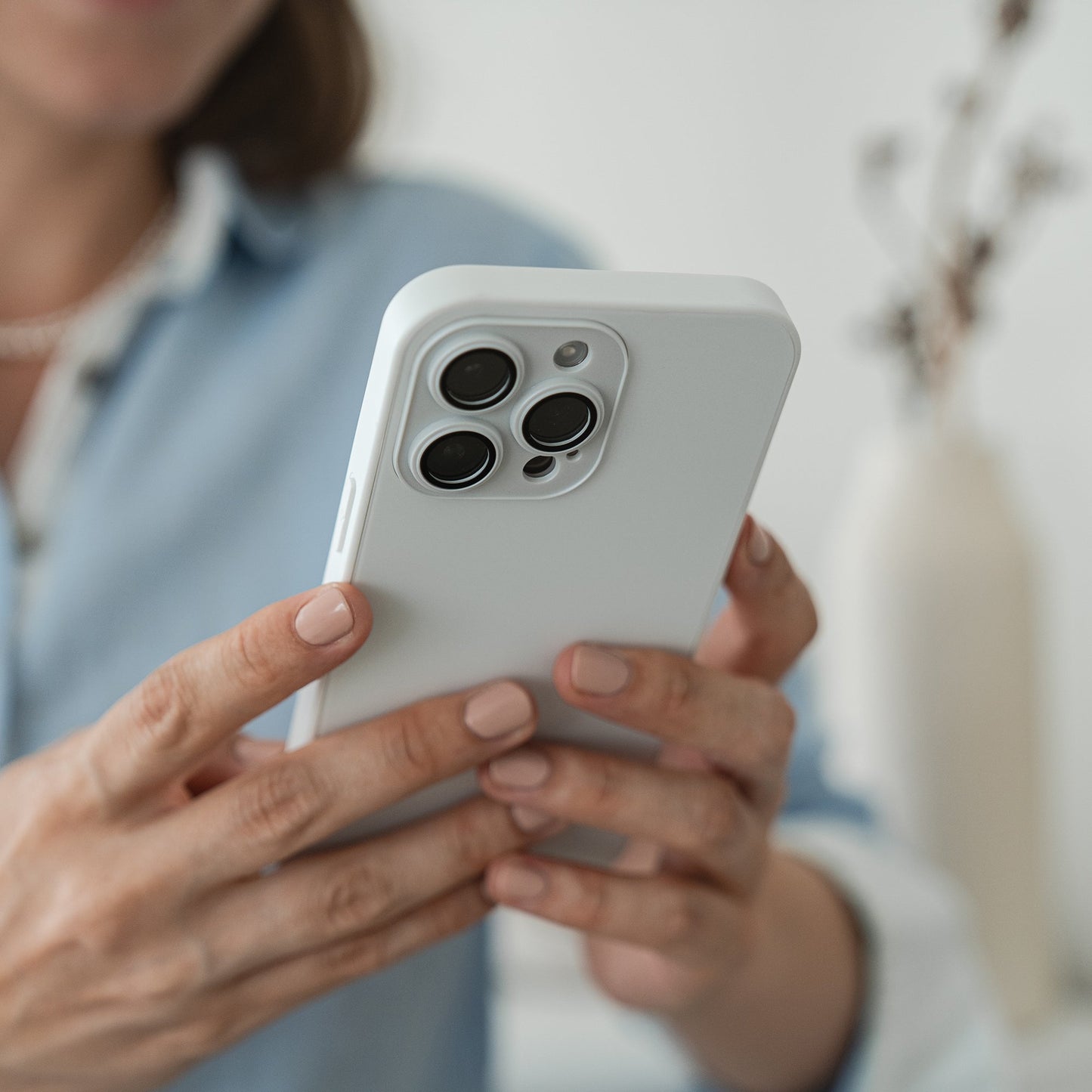 Eine Person in hellblauem Shirt hält das arktis iPhone 16 Tech Case aus stoßabsorbierendem TPU-Silikon von arktis in Weiß mit drei Kameralinsen. Im Hintergrund sind eine weiße Vase und getrocknete Blumen unscharf zu sehen.