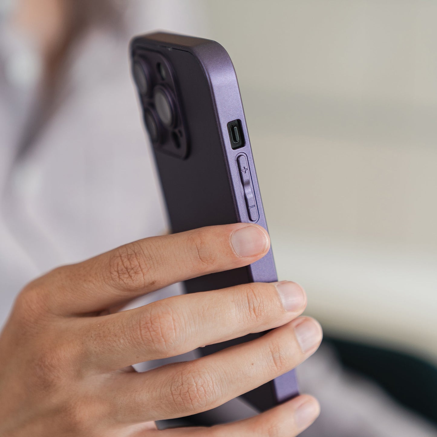 Jemand hält ein lila Smartphone mit dem arktis iPhone 16 Tech Case von arktis aus stoßabsorbierendem TPU-Silikon in der linken Hand, auf dem die seitlichen Tasten und der Kamerabuckel vor einem leicht unscharfen Hintergrund zu sehen sind.