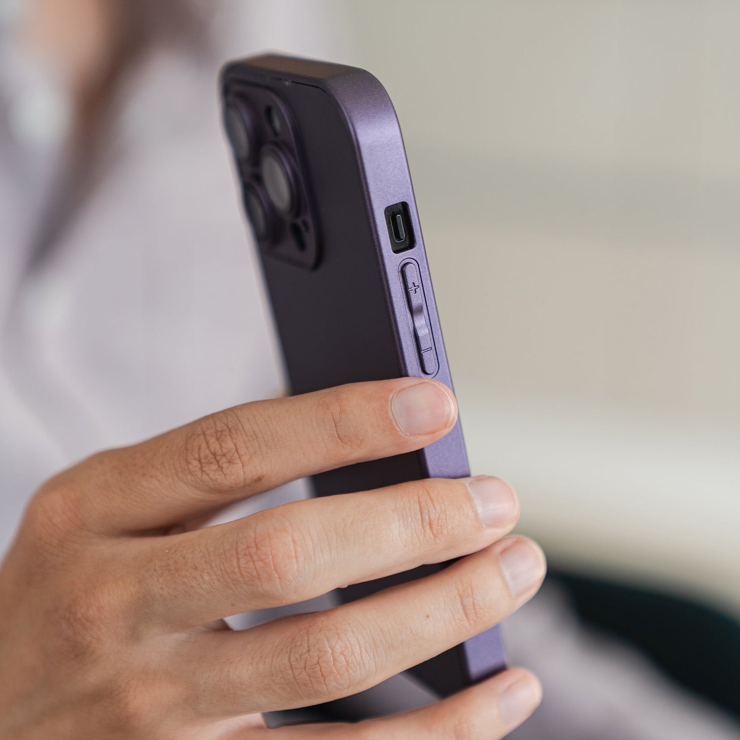 Jemand hält ein lila Smartphone mit dem arktis iPhone 16 Tech Case von arktis aus stoßabsorbierendem TPU-Silikon in der linken Hand, auf dem die seitlichen Tasten und der Kamerabuckel vor einem leicht unscharfen Hintergrund zu sehen sind.
