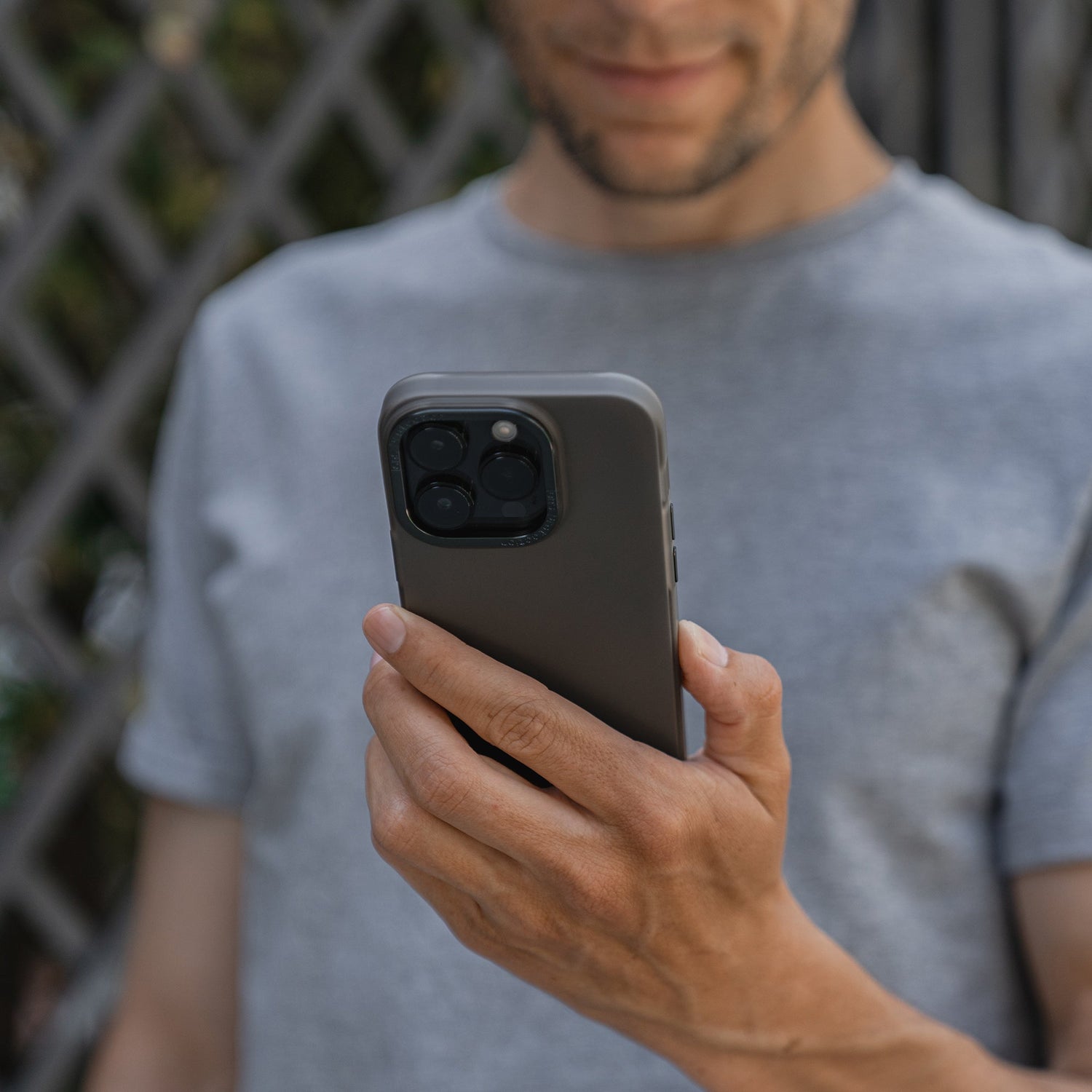 Mann in einem grauen T-Shirt im Freien hält sein Smartphone in der Hand und schaut darauf. Es ist mit der MR PROTECT iPhone 15 Plus FROZEN-Hülle ausgestattet – einer robusten, durchscheinenden Schutzhülle aus Polycarbonat von MR PROTECT.