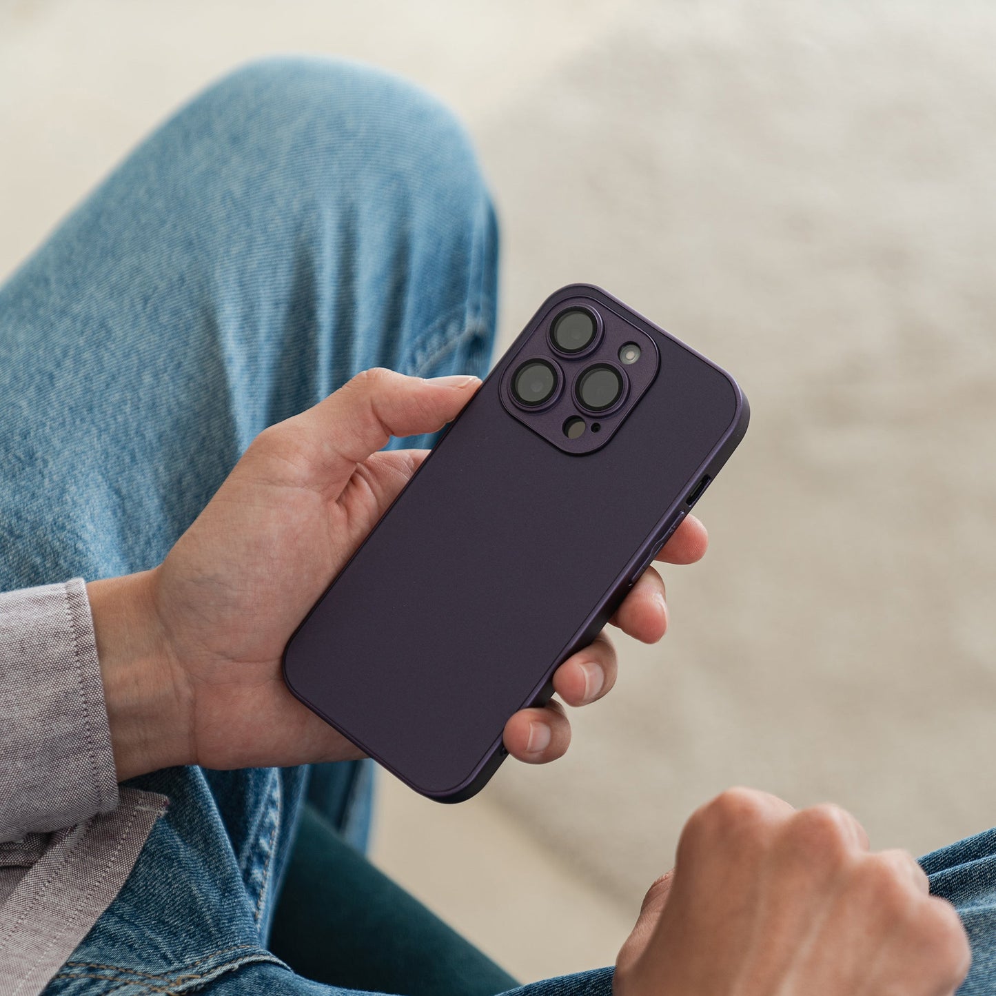 Eine Person in Blue Jeans hält ein Arktis iPhone 16 Tech Case von Arktis in Dunkelviolett und zeigt im Sitzen die Rückseite mit drei Kameraobjektiven vor unscharfem Hintergrund.