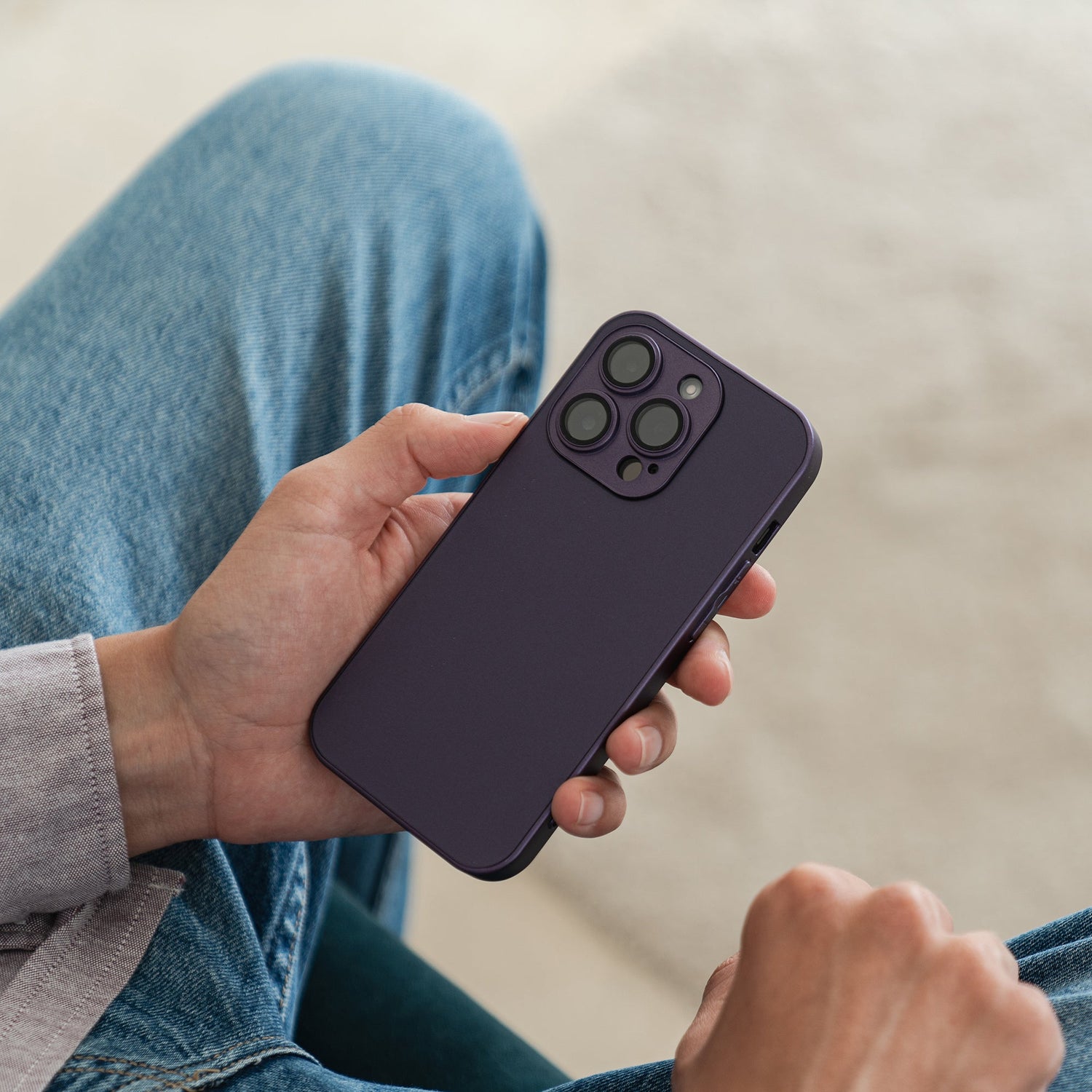 Eine Person in Blue Jeans hält ein Arktis iPhone 16 Tech Case von Arktis in Dunkelviolett und zeigt im Sitzen die Rückseite mit drei Kameraobjektiven vor unscharfem Hintergrund.